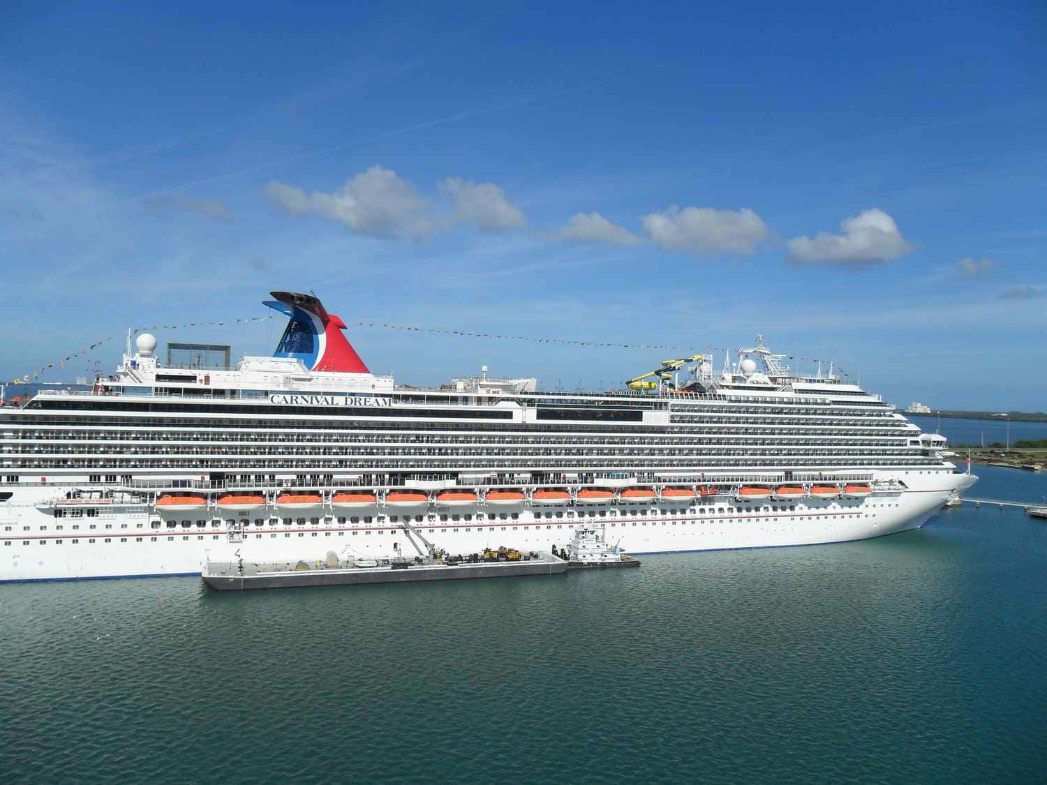 Carnival Dream 桌布 壁紙 — 嘉年華夢想號郵輪在明亮的藍天和散落的雲朵下停泊，白色船體和橙色救生艇與平靜的碧綠海水形成對比。