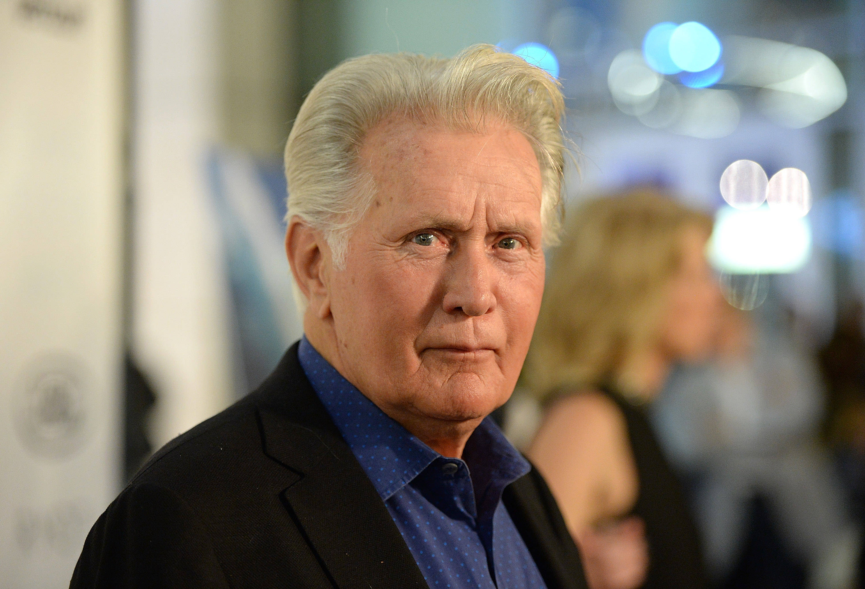 actor 桌布 壁紙 — 嚴肅白髮老年男子特寫肖像，身穿黑色西裝外套和藍色襯衫，背景為模糊活動場景。