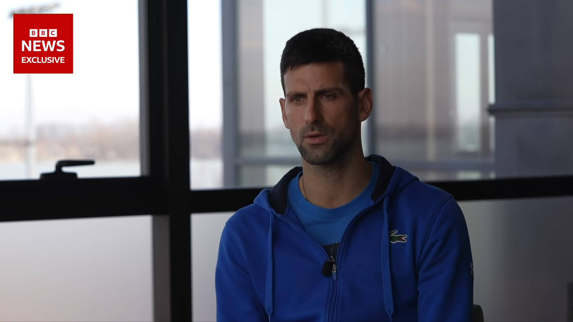 Novak 桌布 壁紙 — 嚴肅的網球選手諾瓦克·喬科維奇穿著藍色連帽衫，在BBC新聞獨家專訪期間坐在室內大窗戶附近。