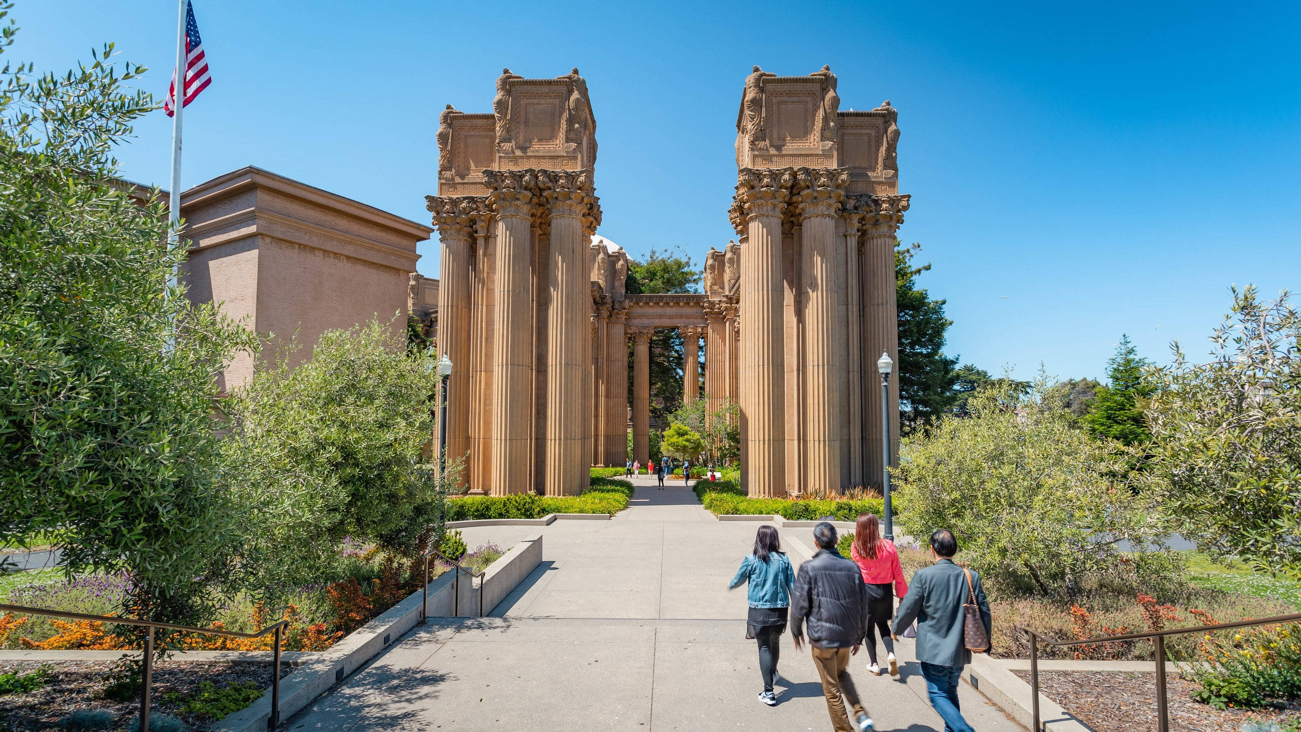 人物 桌布 壁紙 — 四人在陽光普照的小徑上漫步，走向宏偉的柱廊式 Palace of Fine Arts，天空湛藍清澈。