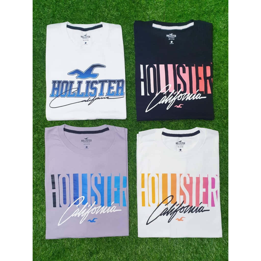Hollister 桌布 壁紙 — 四件折疊好的Hollister California T恤，顏色分別為白色、海軍藍、薰衣草紫和白色，帶有彩色標誌，展示在綠色草地上。