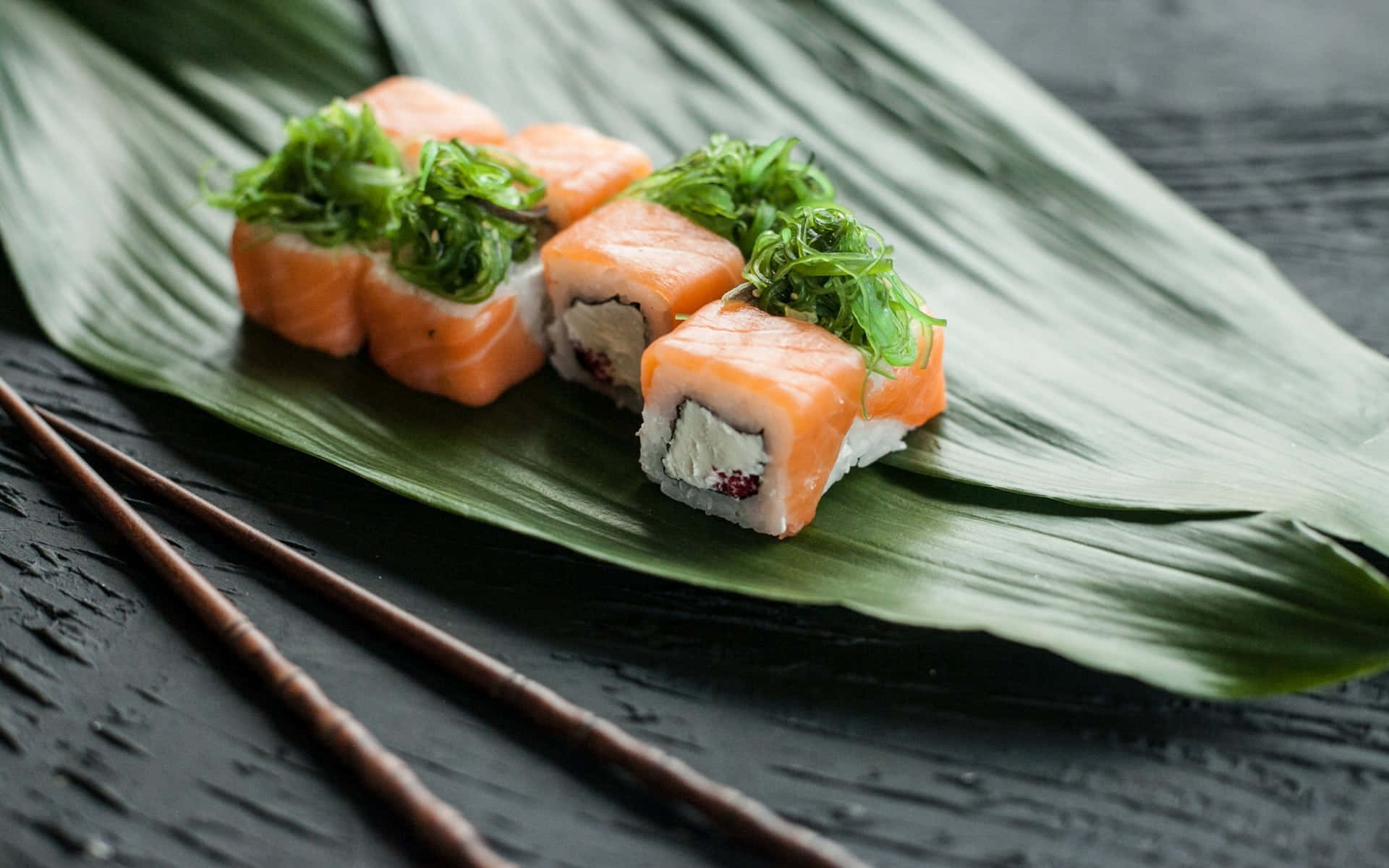 Types Of Sushi 桌布 壁紙 — 四件鮭魚壽司卷配上新鮮蔬菜，放在深色木質表面上的綠葉上，旁邊有筷子。