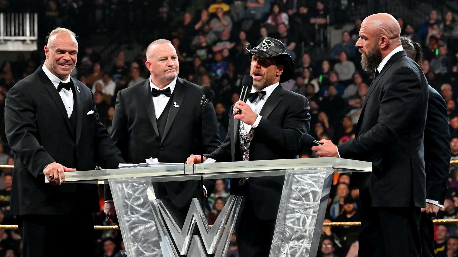 WWE 桌布 壁紙 — 四位WWE傳奇人物身穿燕尾服，站在印有WWE標誌的玻璃講台上，在擠滿觀眾的場館內進行摔角活動。