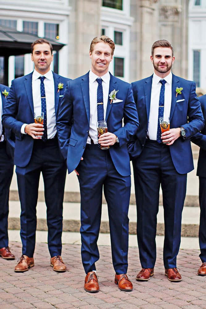 Groomsmen 桌布 壁紙 — 四位伴郎穿著相配的海軍藍西裝和棕色鞋子，站在石頭庭院上微笑著手持啤酒杯。