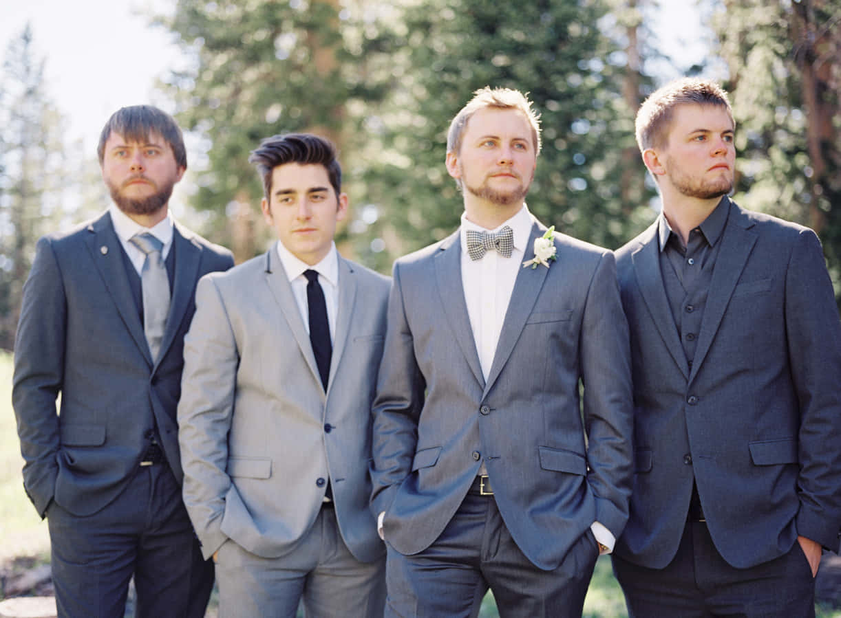 Groomsmen 桌布 壁紙 — 四位伴郎穿著量身訂做的西裝，在明亮的陽光下自信地站在戶外，身後有樹木。