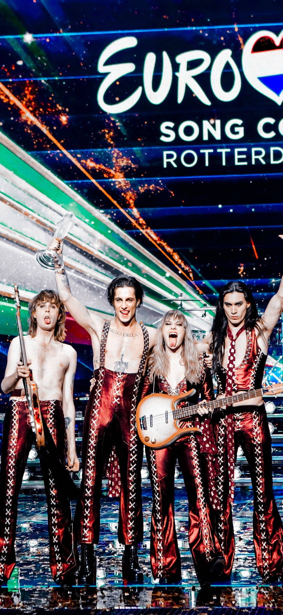 Maneskin 桌布 壁紙 — 四位充滿活力的表演者身穿閃亮紅色服裝，在閃閃發光的歐洲歌唱大賽舞台上，手持獎盃和吉他慶祝。