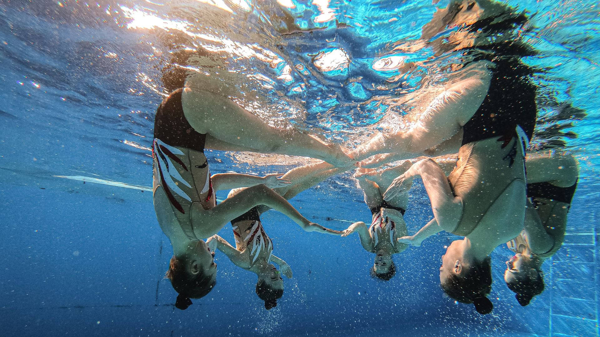 Swimming 桌布 壁紙 — 四位同步泳者在水下形成一個圓圈，陽光穿過水面過濾。