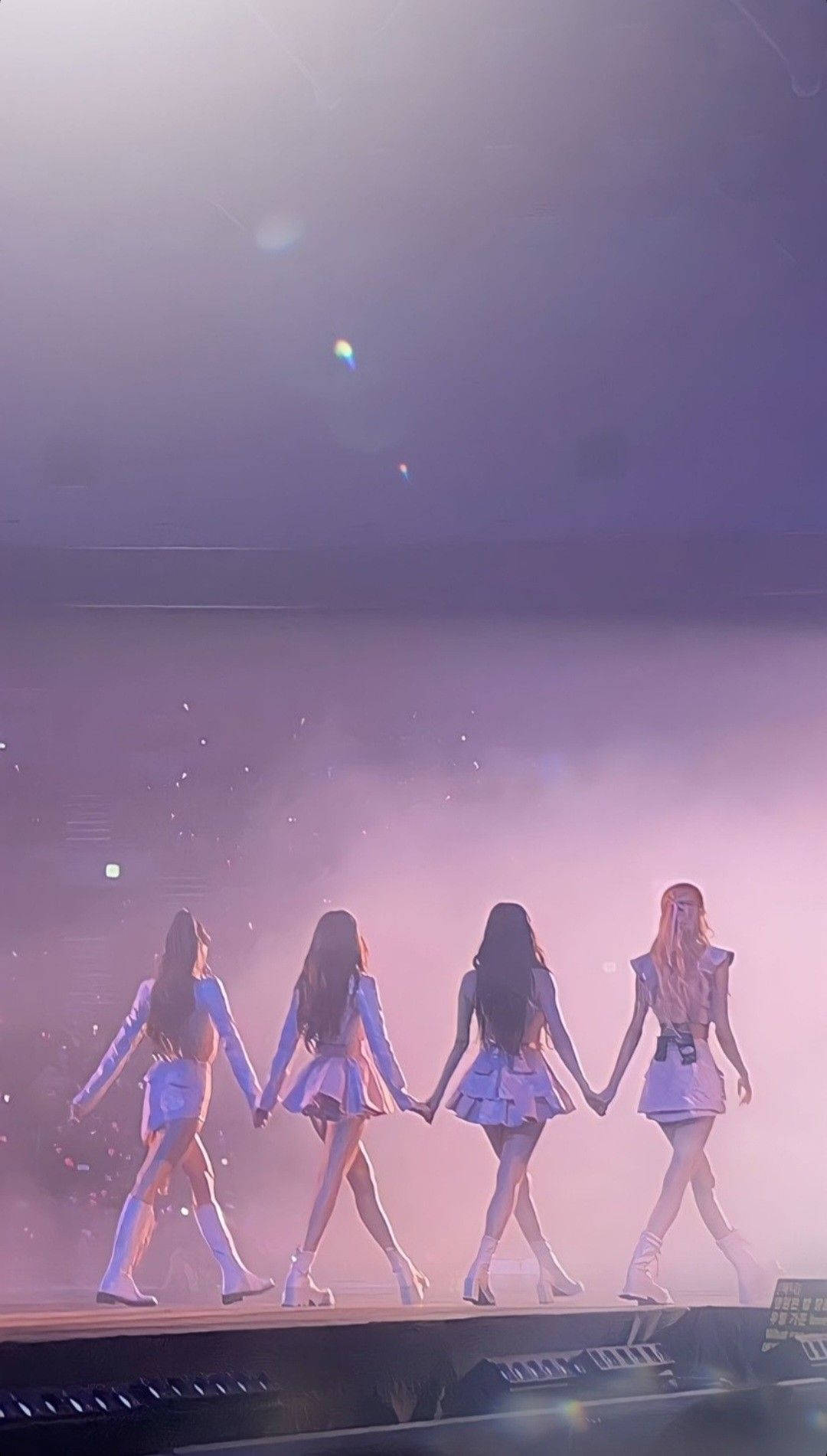 BLACKPINK 桌布 壁紙 — 四位女表演者手牽手走過一個煙霧瀰漫、沐浴在紫色光線和鏡頭光暈中的舞台。