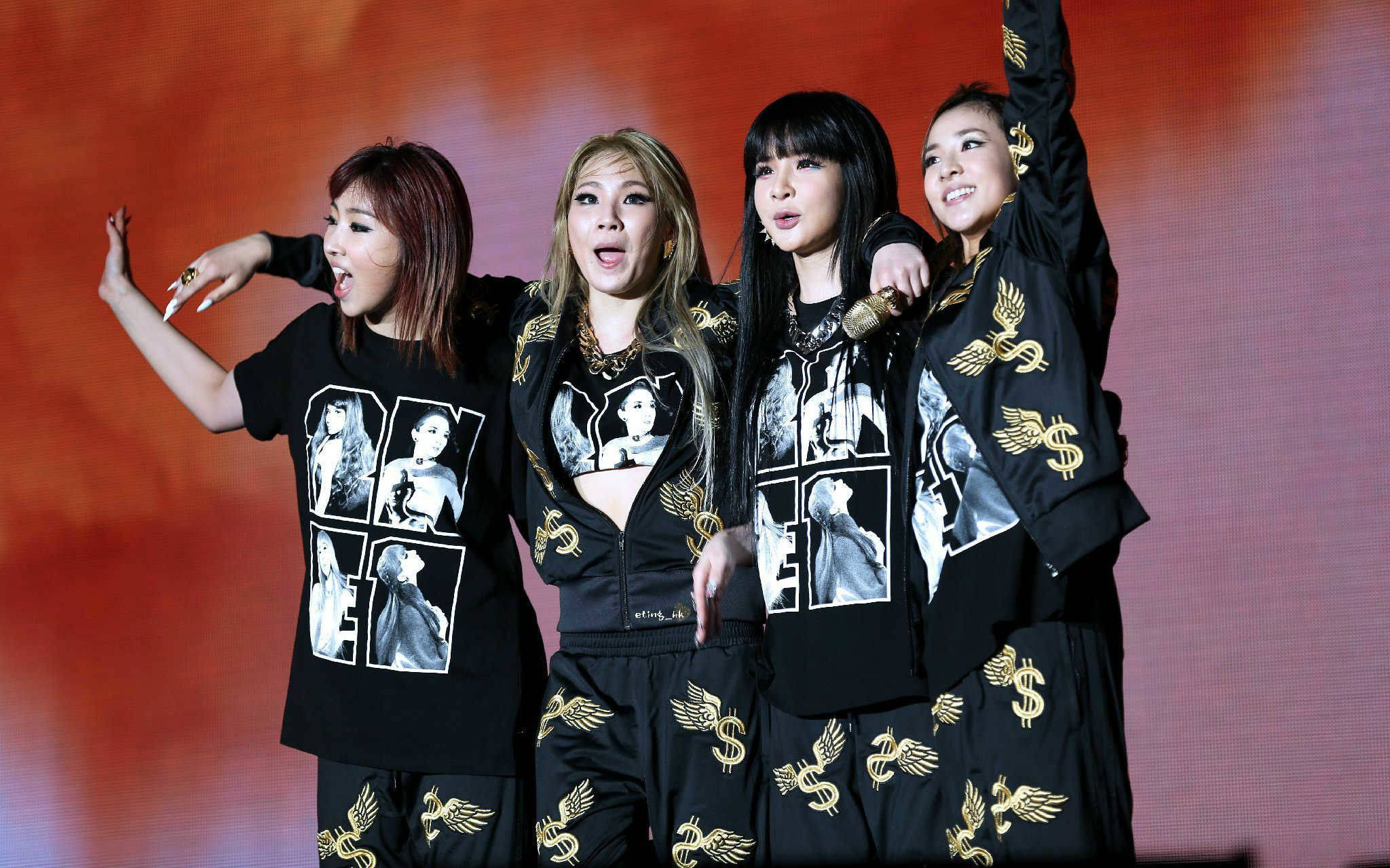2ne1 桌布 壁紙 — 四位女表演者身穿黑色服裝，飾有金色美元符號和帶翅膀的標誌，在發光的橙色舞台上擺出動態姿勢。