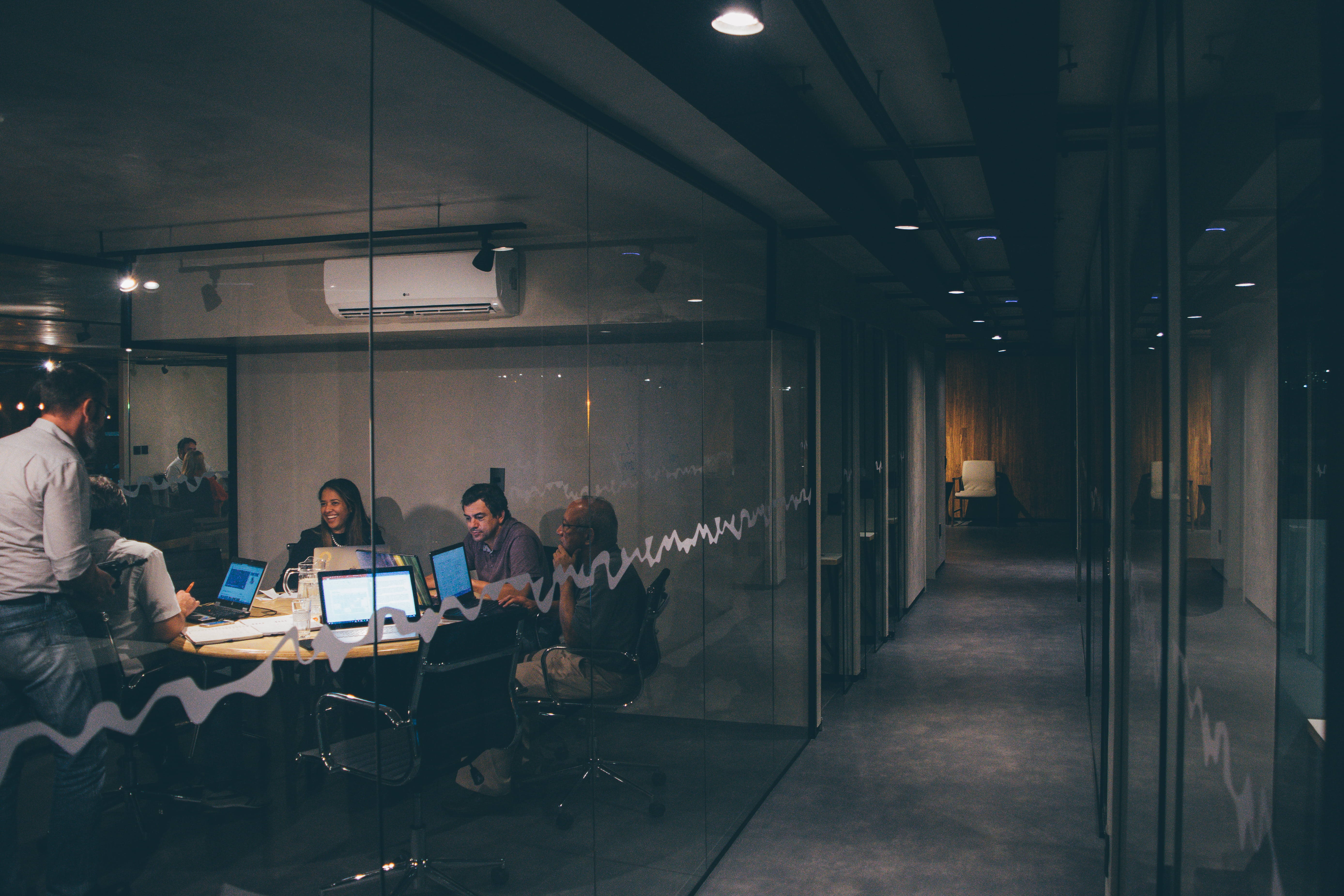 Working 桌布 壁紙 — 四位專業人士在光線昏暗的傍晚，於現代玻璃牆辦公室內專注地使用筆記型電腦工作。