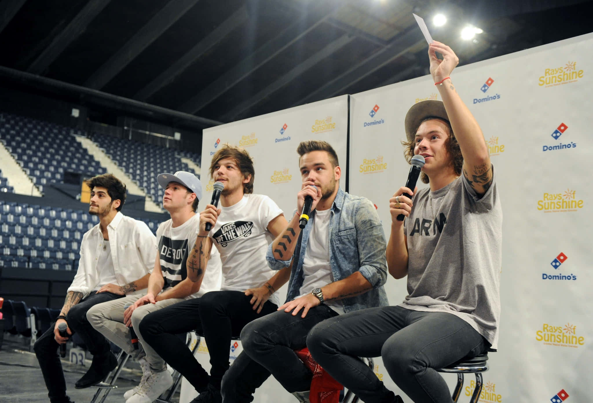 One Direction 桌布 壁紙 — 四位年輕男性坐在舞台上的凳子上，手持麥克風，在一個後面有空的藍色座位的場館中進行問答活動。