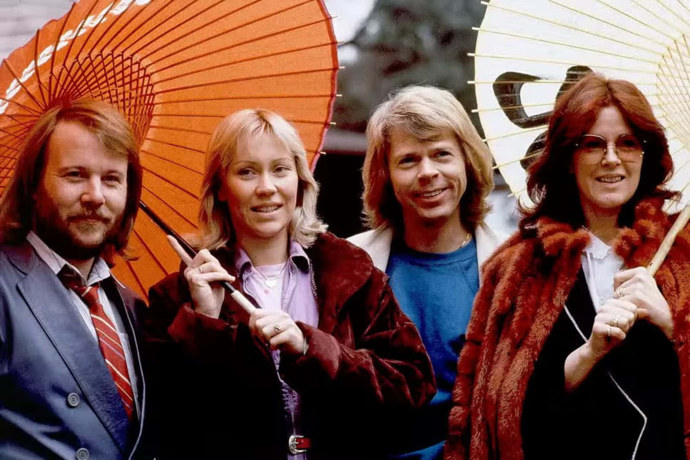 阿巴 桌布 壁紙 — 四位微笑的1970年代音樂家手持華麗紙傘，在模糊的戶外背景下擺姿勢。
