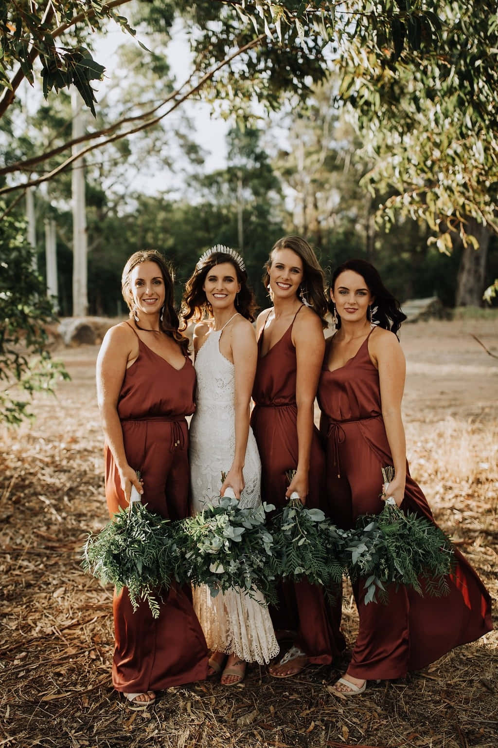 Bridesmaids 桌布 壁紙 — 四位微笑的女性，身穿鏽色連衣裙和白色婚紗，一起站在枝葉茂密的樹下，手持綠色花束。