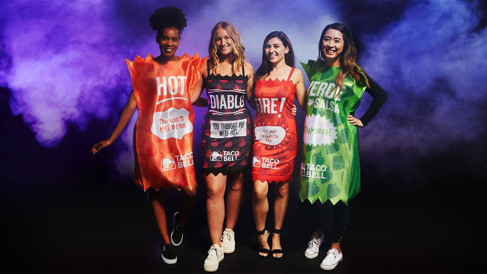 Taco Bell 桌布 壁紙 — 四位微笑的女性穿著Taco Bell醬料服裝，在煙霧繚繞的紫色和藍色背景前擺姿勢。
