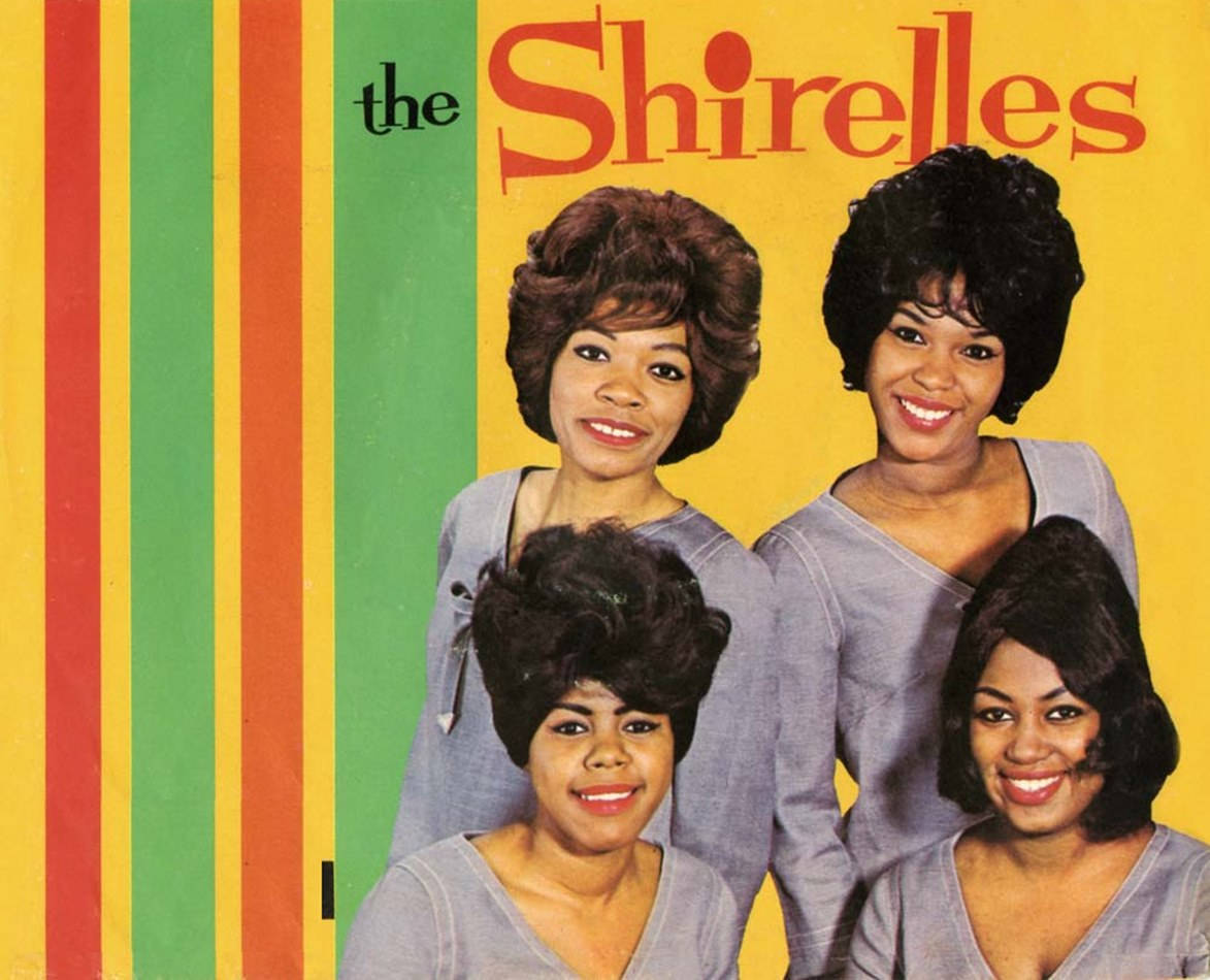 Album Cover 桌布 壁紙 — 四位微笑的女性穿著相配的灰色上衣，在充滿活力的黃、綠、紅條紋背景前擺姿勢，背景上有粗體紅色的「the Shirelles」字樣。