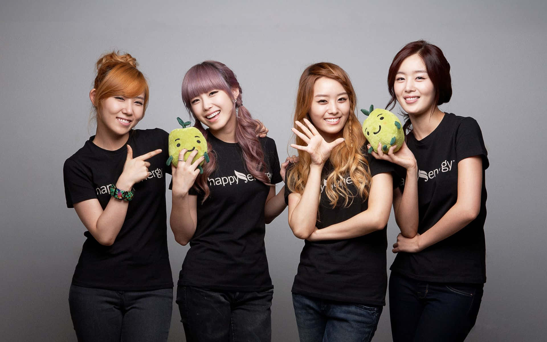 K-pop 桌布 壁紙 — 四位微笑的年輕女性穿著黑色T恤，在純灰色工作室背景前愉快地拿著綠色毛絨蘋果擺姿勢。