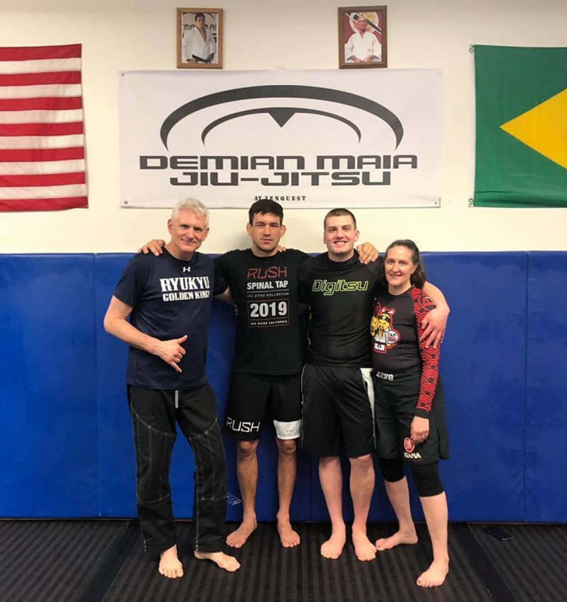 Champion 桌布 壁紙 — 四位微笑的武術家在鋪有藍色軟墊和掛有Demian Maia柔術橫幅的健身房中一起擺姿勢。