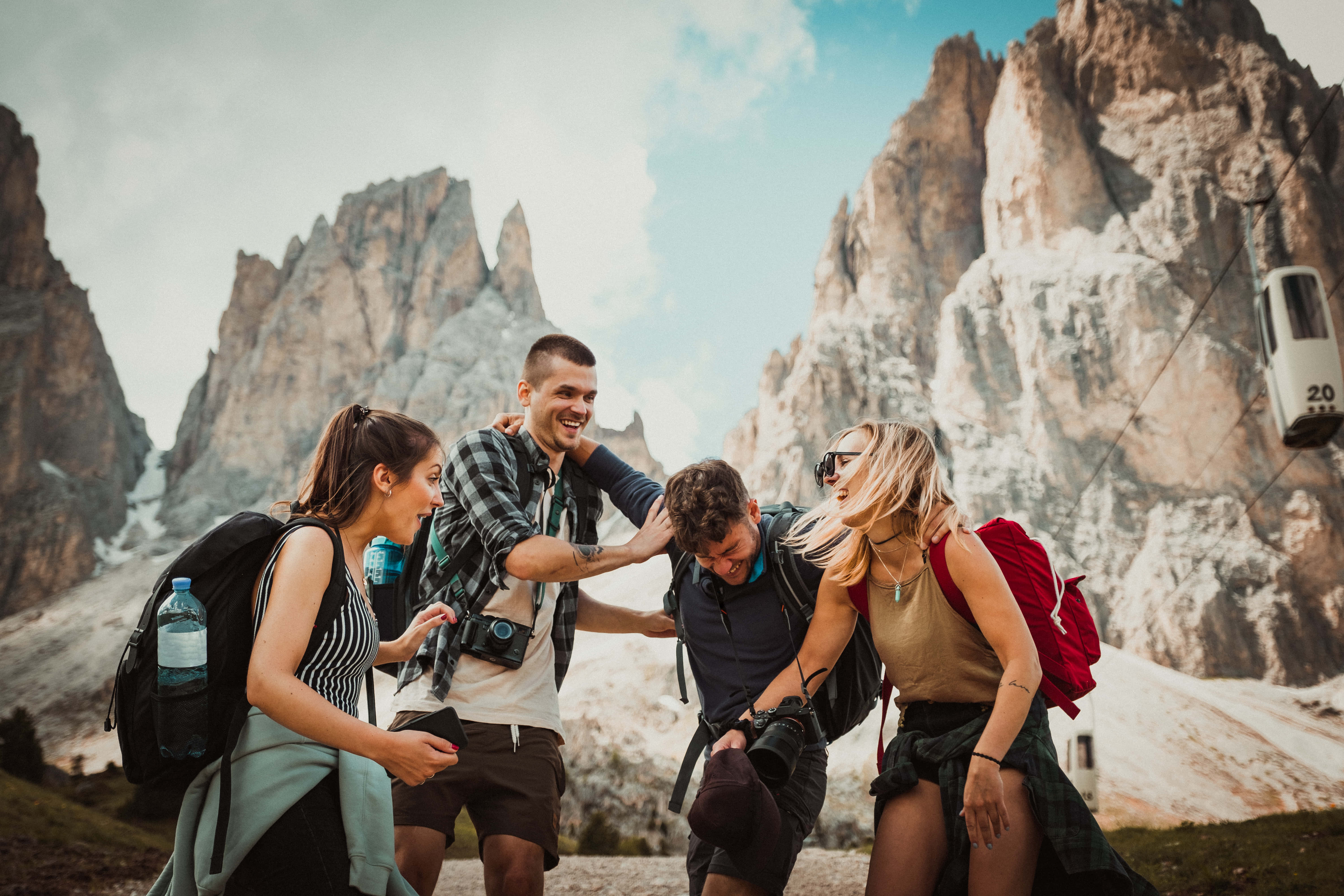 World Travel 桌布 壁紙 — 四位微笑的遠足者揹著背包，在崎嶇的山峰和纜車背景下，於山徑上一起大笑。