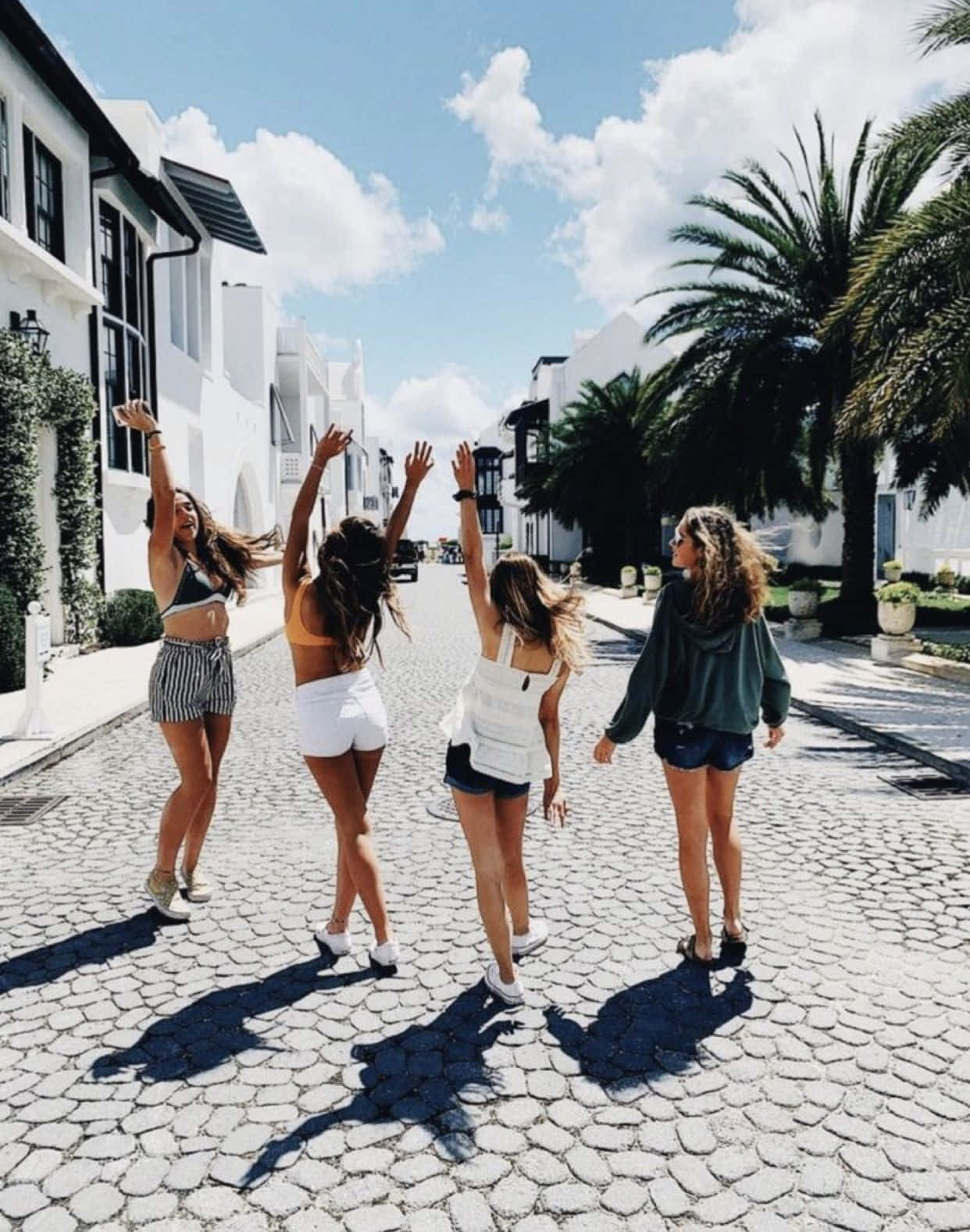 girls 桌布 壁紙 — 四位快樂的朋友在慶祝中舉起手臂，沿著一條陽光普照的鵝卵石街道行走，街道兩旁是白色建築和棕櫚樹。