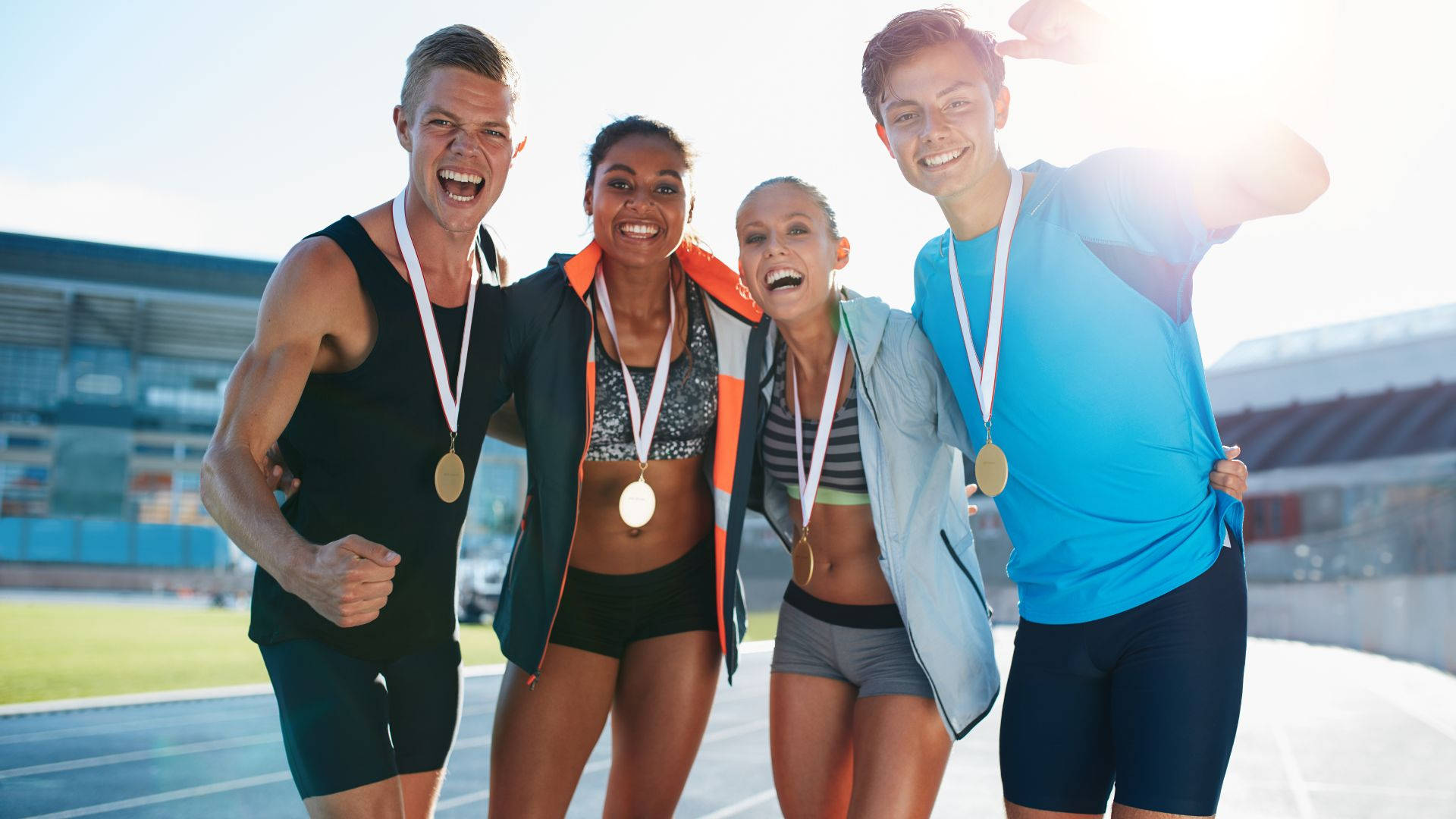 Olympics 桌布 壁紙 — 四位快樂的運動員在明亮的陽光下戴著金牌在跑道上慶祝。