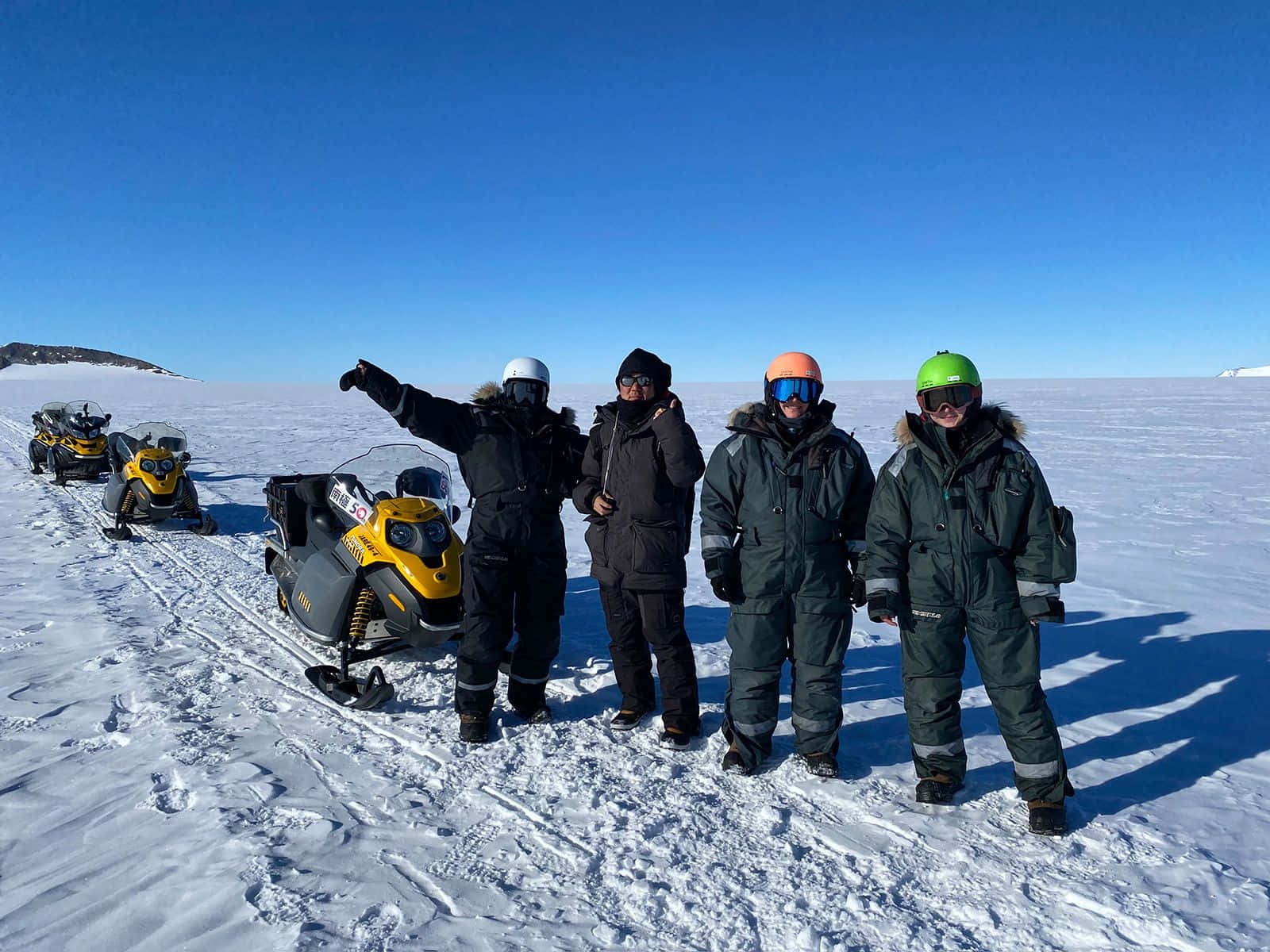 Antarctica 桌布 壁紙 — 四位戴著鮮豔頭盔的雪地摩托車手，在晴朗的藍天下，在一片廣闊的冰凍平原上，自豪地站在他們的黃色機器旁。