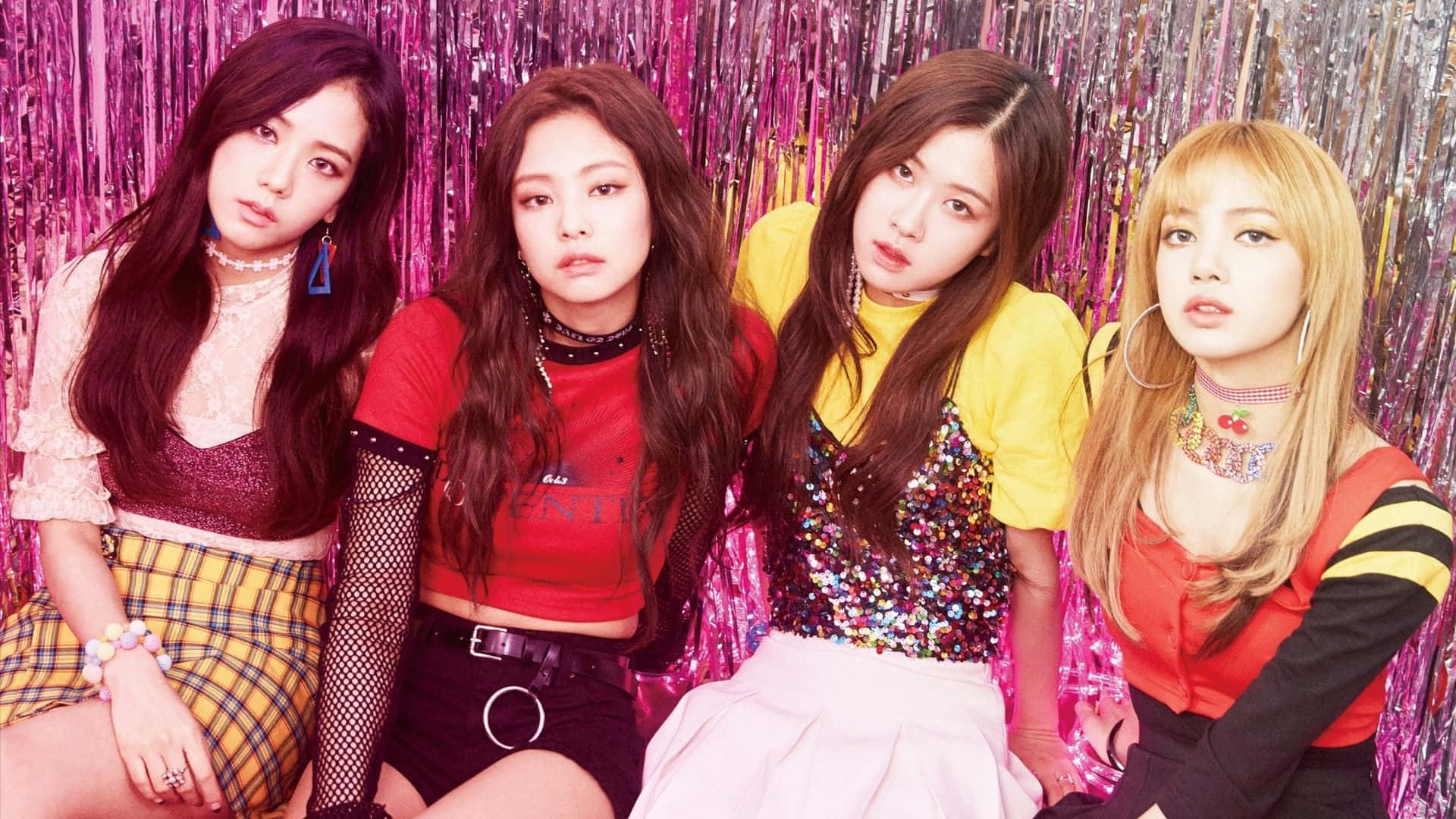 K-pop 桌布 壁紙 — 四位時尚女性在閃爍的粉紅色金屬箔背景前，穿著大膽多彩的服裝自信地擺姿勢。