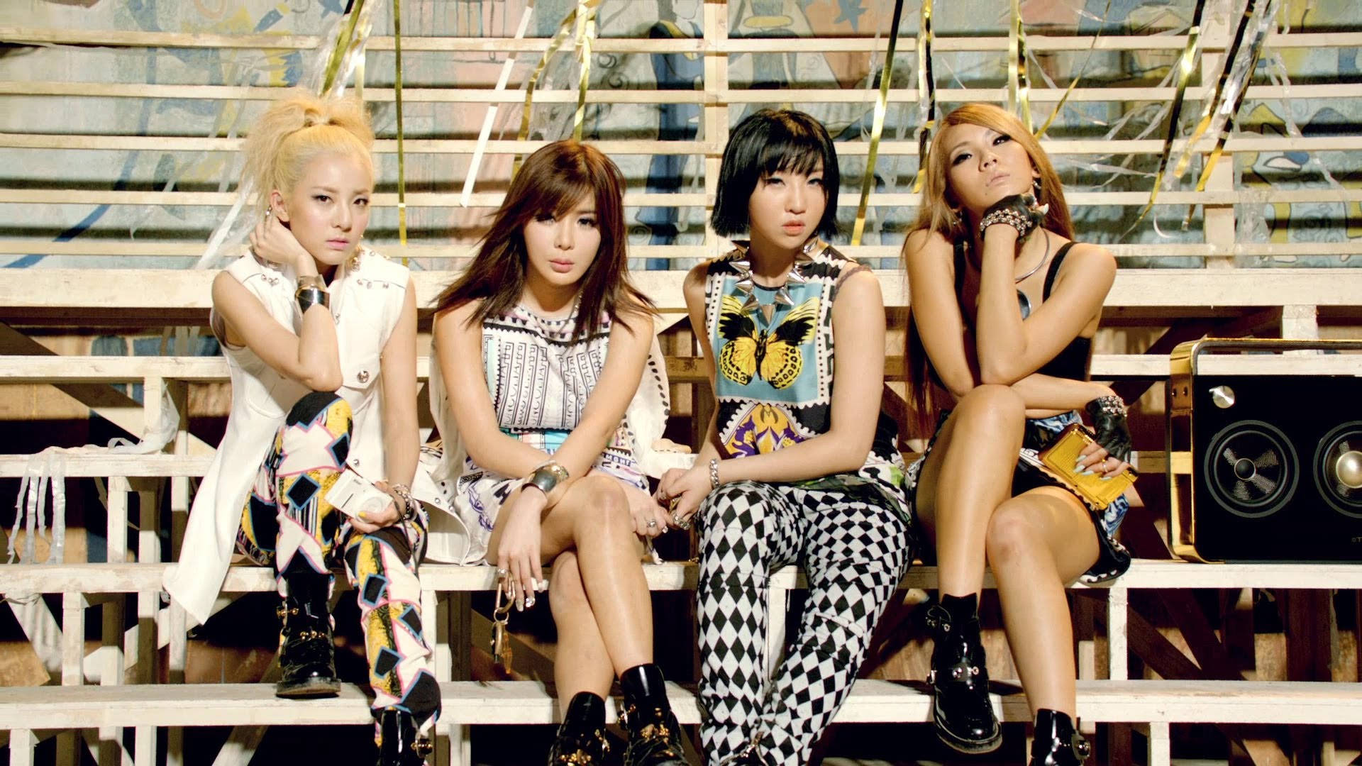 2ne1 桌布 壁紙 — 四位時尚女性穿著大膽圖案服裝，在風化的看台上自信擺姿勢，背景有塗鴉和揚聲器。