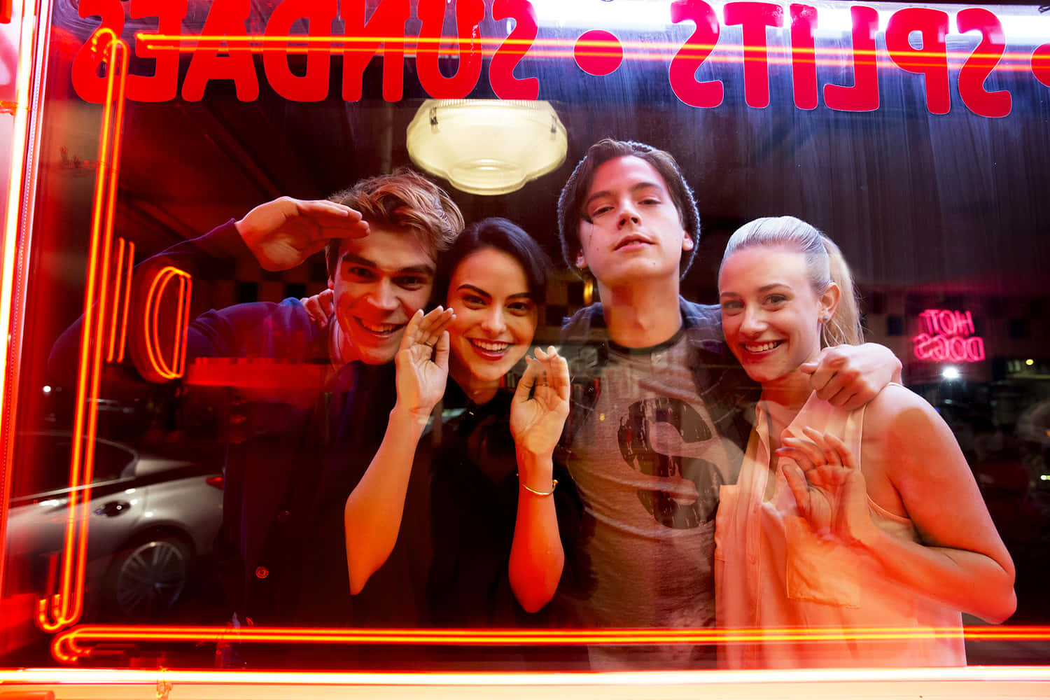 Riverdale 桌布 壁紙 — 四位朋友在夜晚於發光的紅色霓虹燈窗後愉快地擺姿勢，微笑並做出俏皮的手勢。