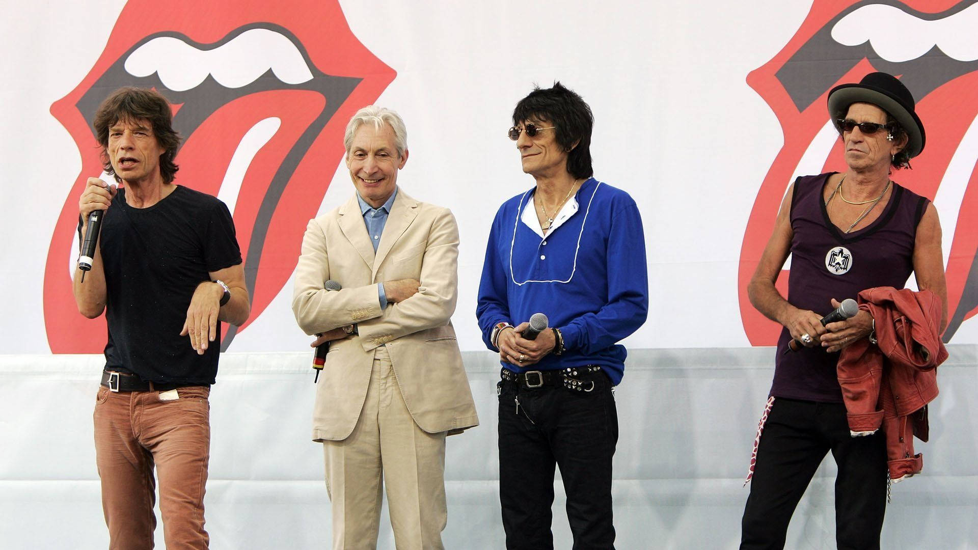 Rock 桌布 壁紙 — 四位標誌性搖滾音樂家站在舞台上，背景是巨大的滾石樂隊舌頭標誌。