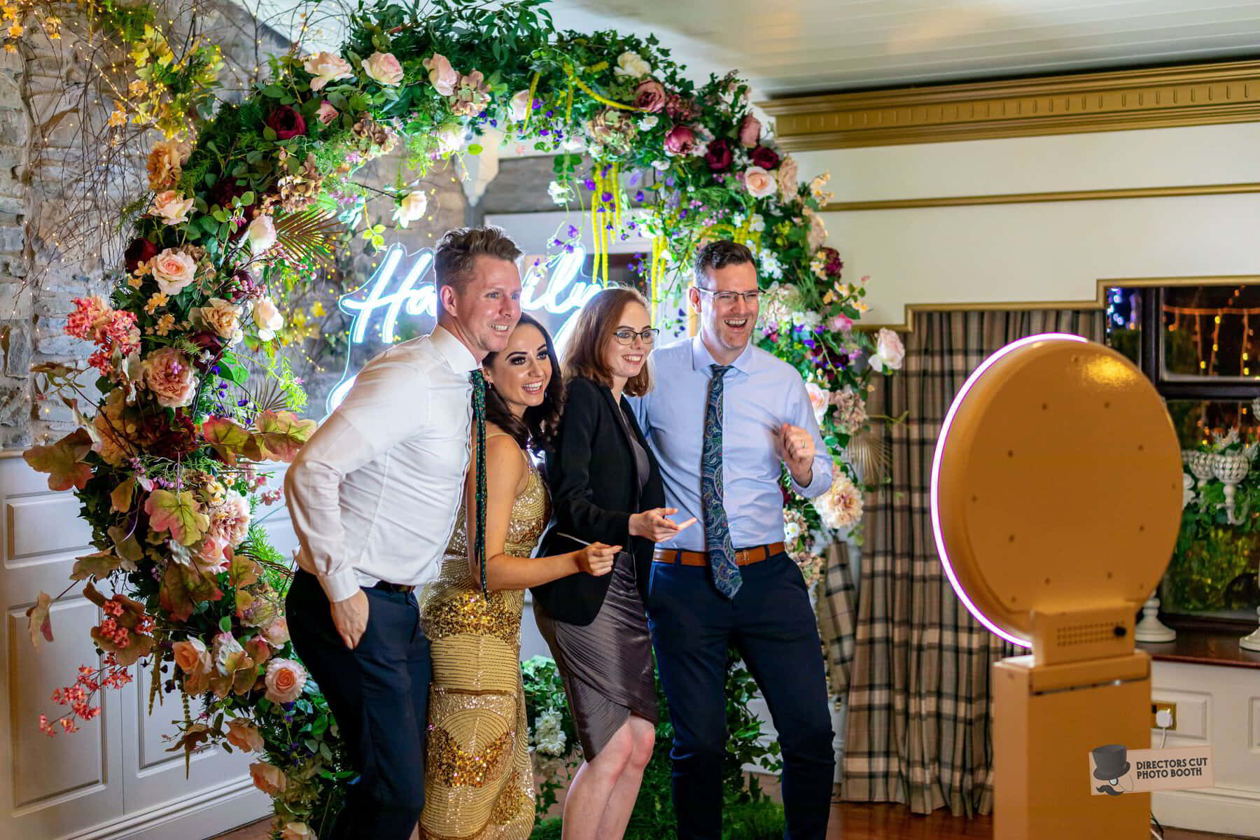 Photo Booth 桌布 壁紙 — 四位歡樂的賓客在一個充滿活力的花卉拱門前擺姿勢，拱門上有一個發光的霓虹燈標誌，背景是節日室內活動。