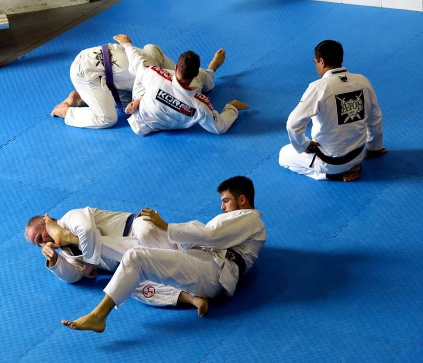 martial arts 桌布 壁紙 — 四位武術家穿著白色道服，在明亮的藍色墊子上專注地進行巴西柔術訓練。