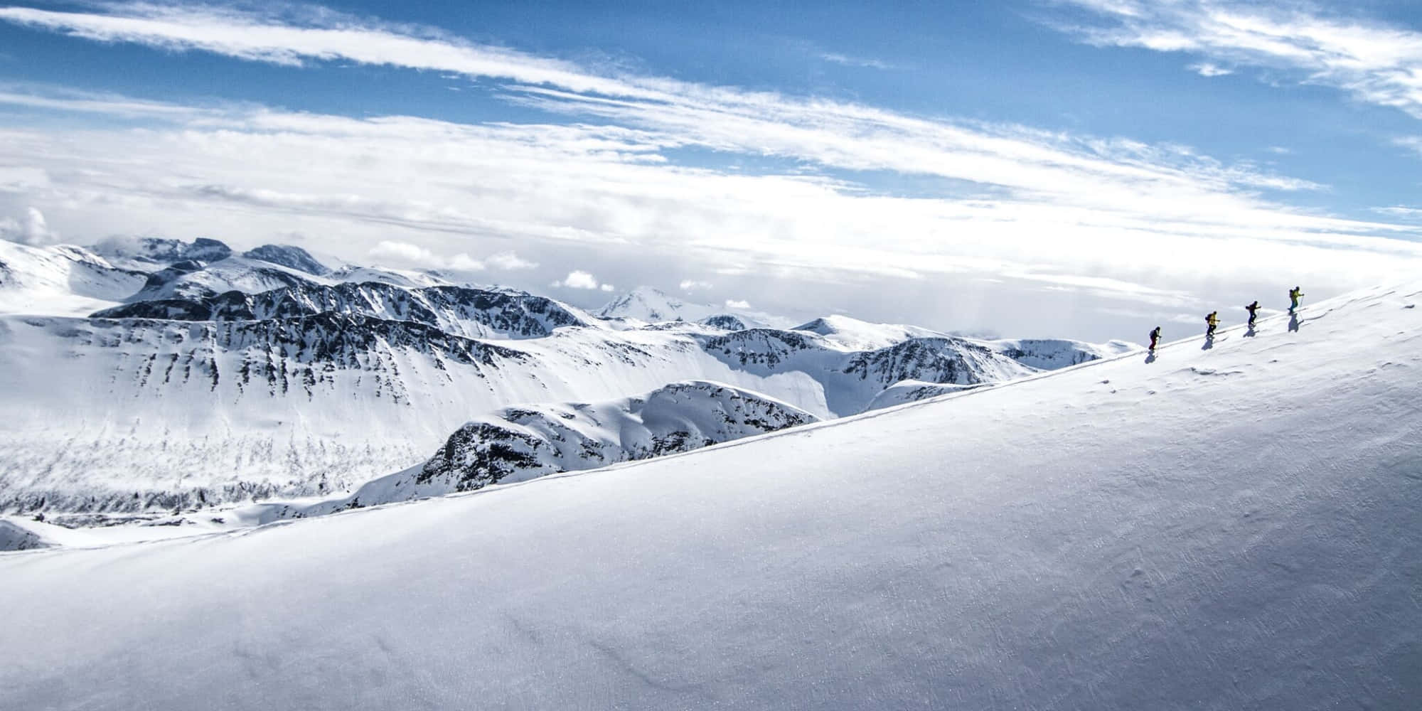 Ski Mountain 桌布 壁紙 — 四位滑雪者在一片廣闊的雪白山脊上攀登，天空湛藍，雲朵輕柔。