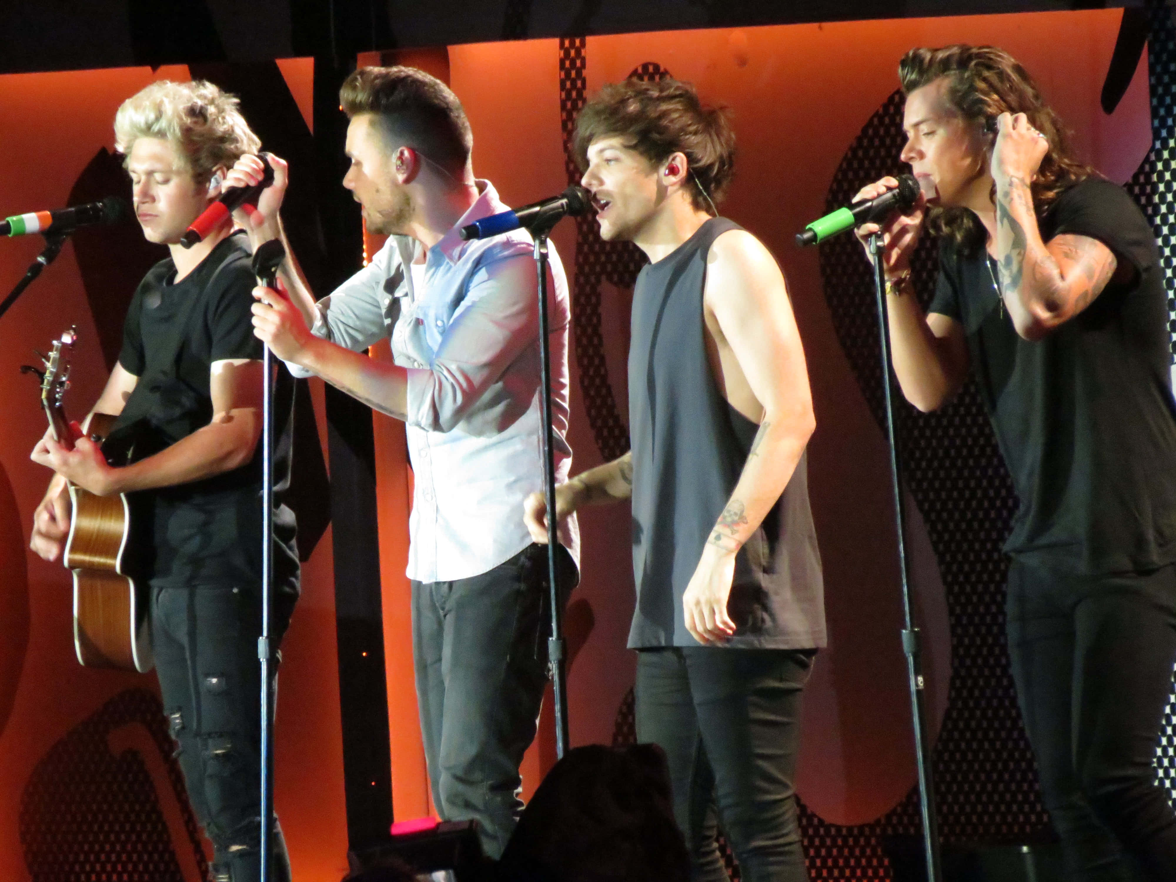 One Direction 桌布 壁紙 — 四位男性樂隊成員在溫暖的橙色燈光下，手持麥克風和吉他，在舞台上充滿活力地表演。