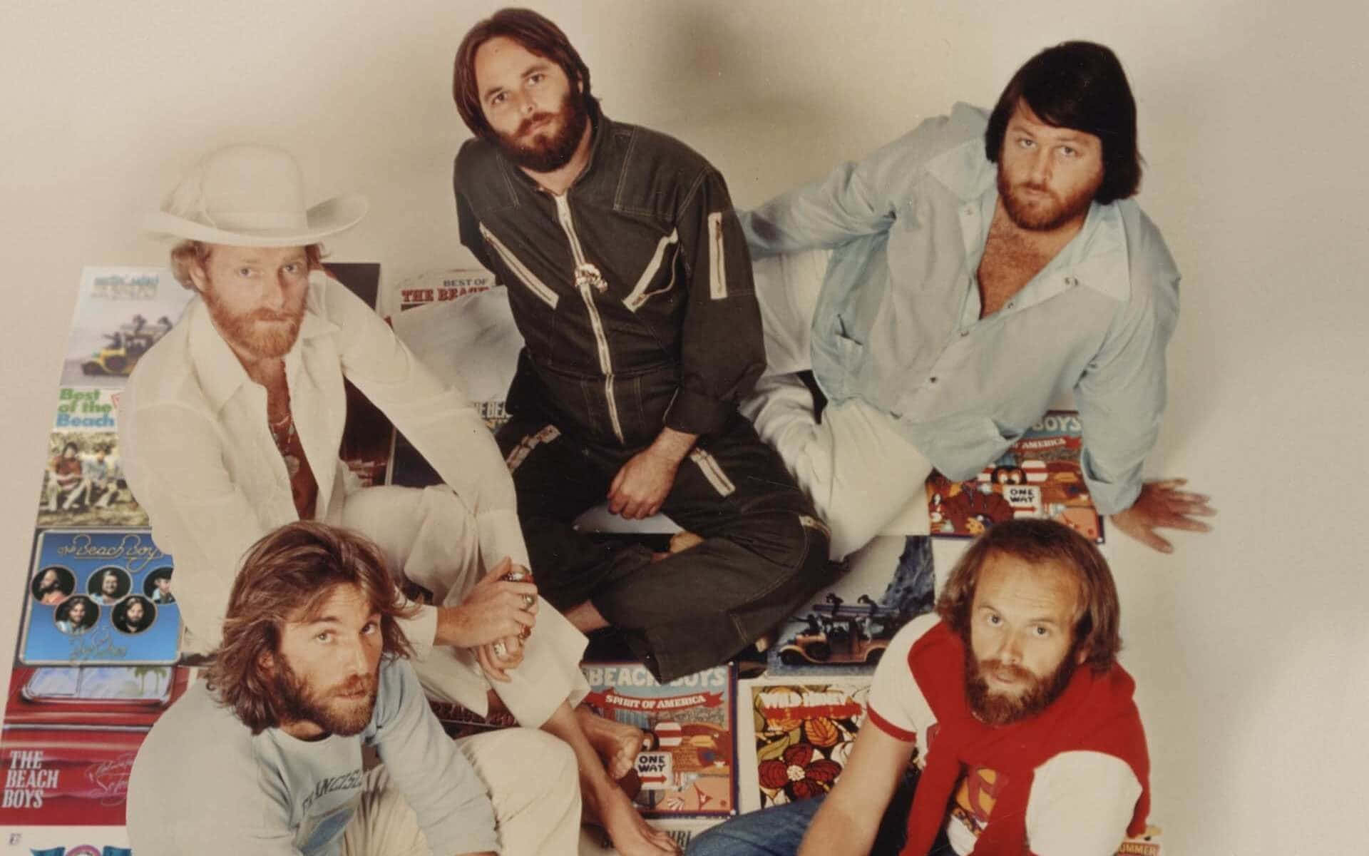 band 桌布 壁紙 — 四位留鬍鬚的男子穿著1970年代的服裝，隨意地擺姿勢坐在一堆 Beach Boys 專輯上，背景是素牆。