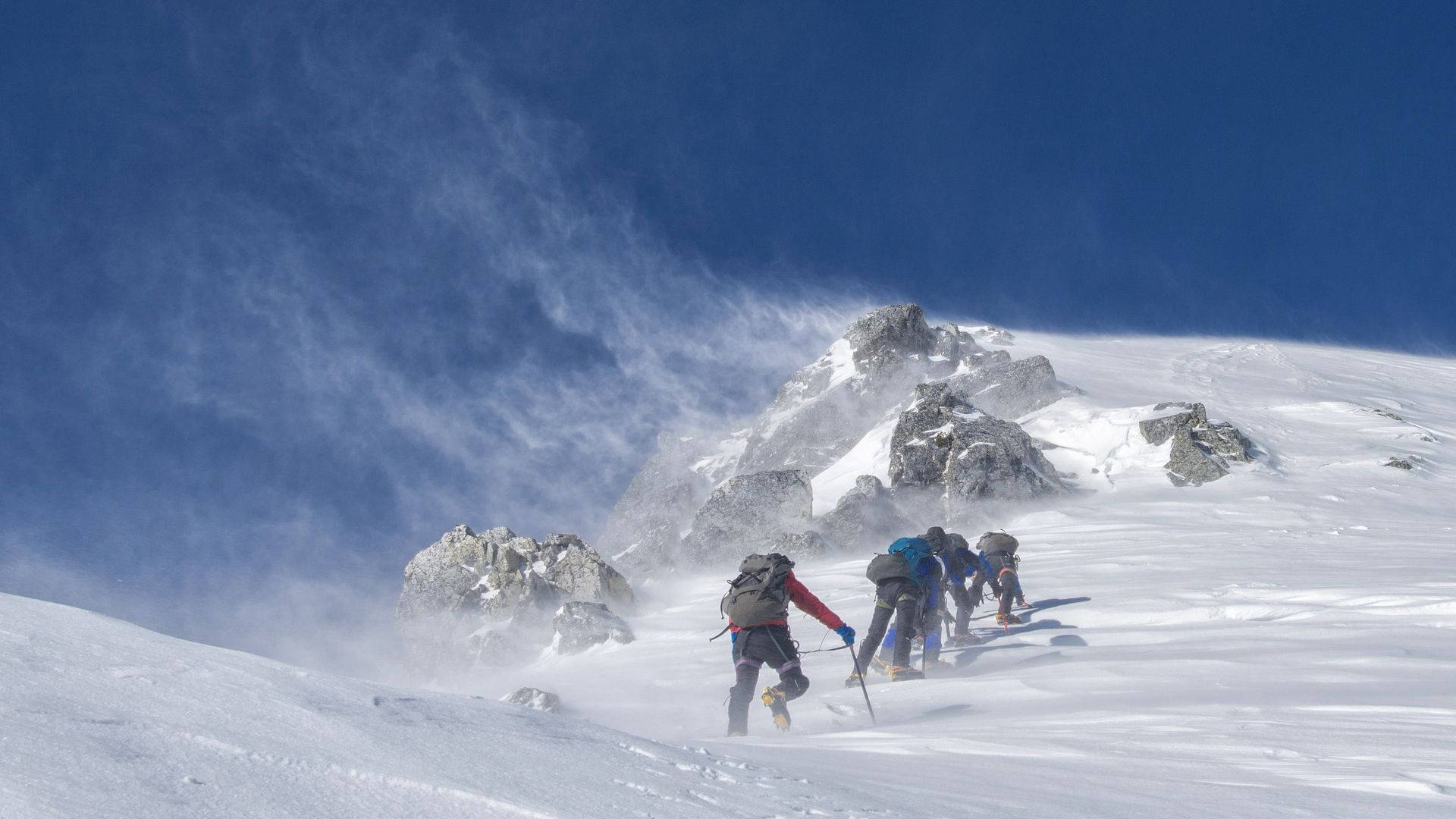 攀爬 桌布 壁紙 — 四位登山者在晴朗的藍天下攀登積雪的山脊，風吹起他們周圍的雪。