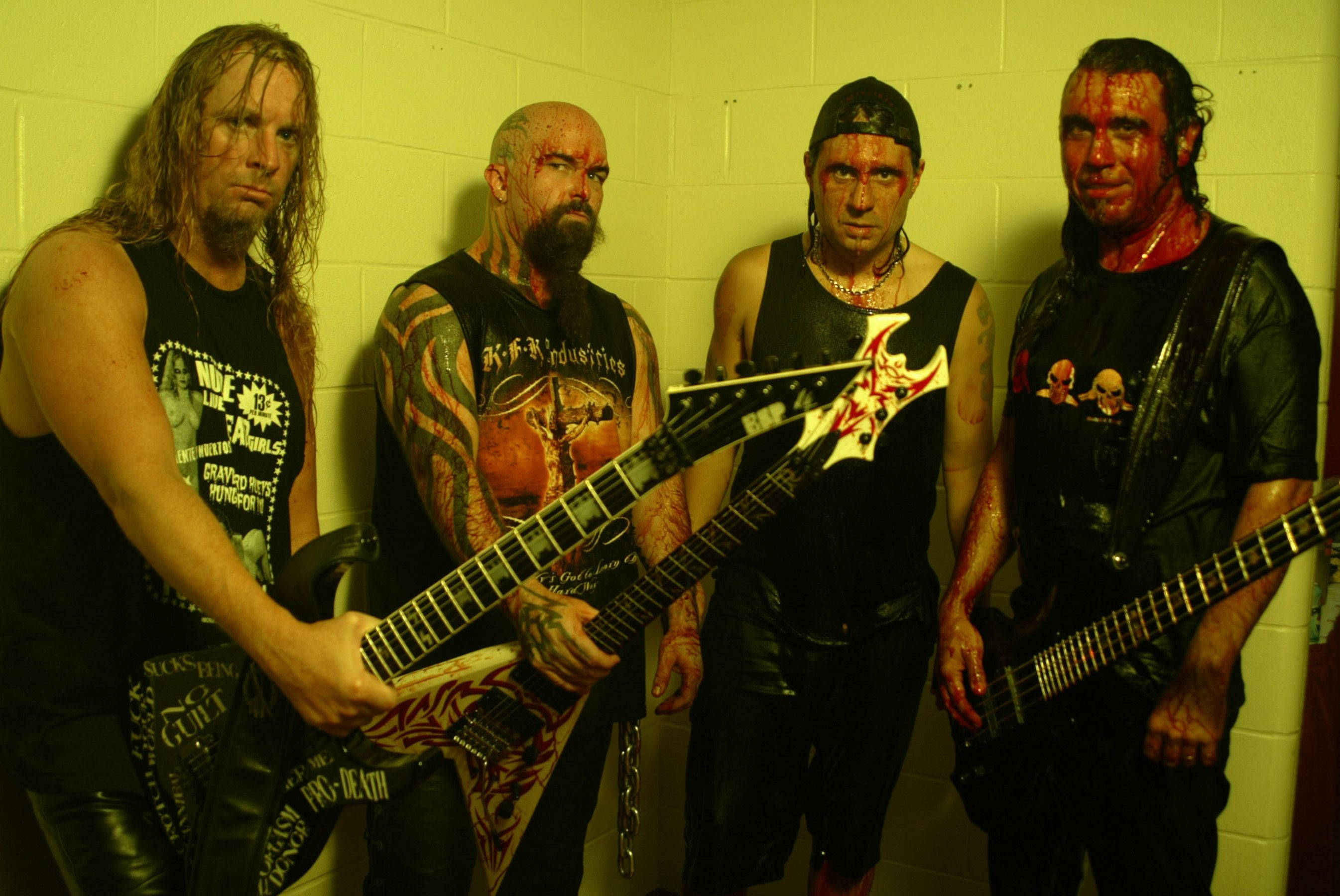 Slayer 桌布 壁紙 — 四位紋身濃重的金屬樂手，滿臉血跡，在黃色調的煤渣磚牆前激烈擺姿勢，手持電吉他。