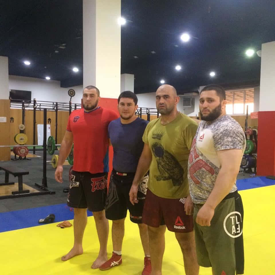 Fitness 桌布 壁紙 — 四位肌肉發達的男士穿著運動服，自信地站在光線充足的健身房內的黃色墊子上，周圍有舉重器材。