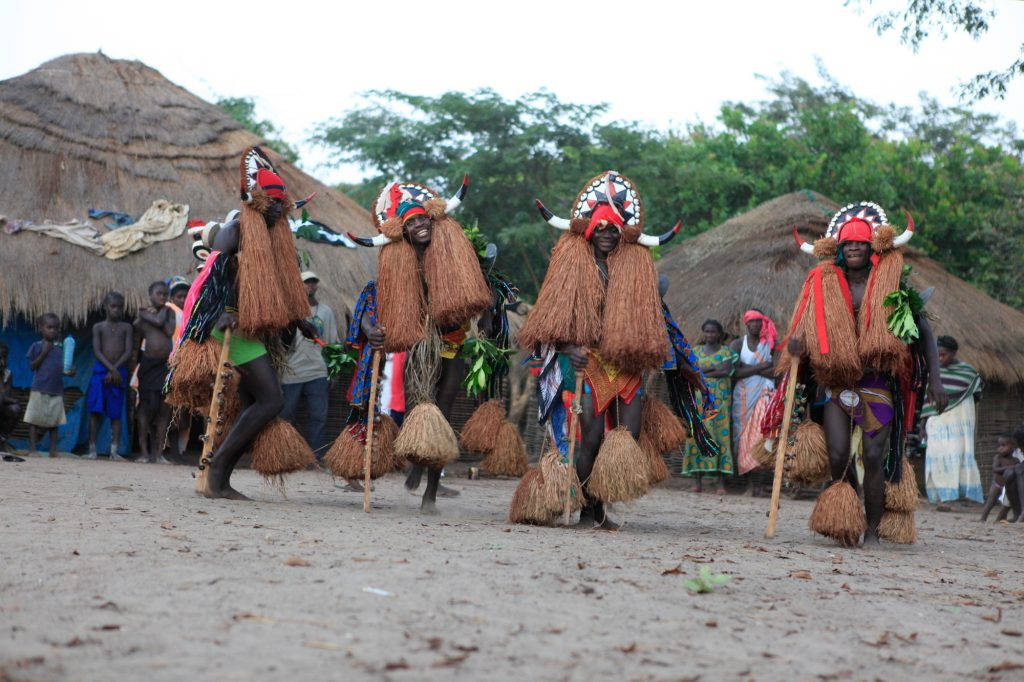 Guinea Bissau 桌布 壁紙 — 四位舞者戴著精緻的稻草和角頭飾，在茅草屋前進行傳統儀式，旁觀者在觀看。