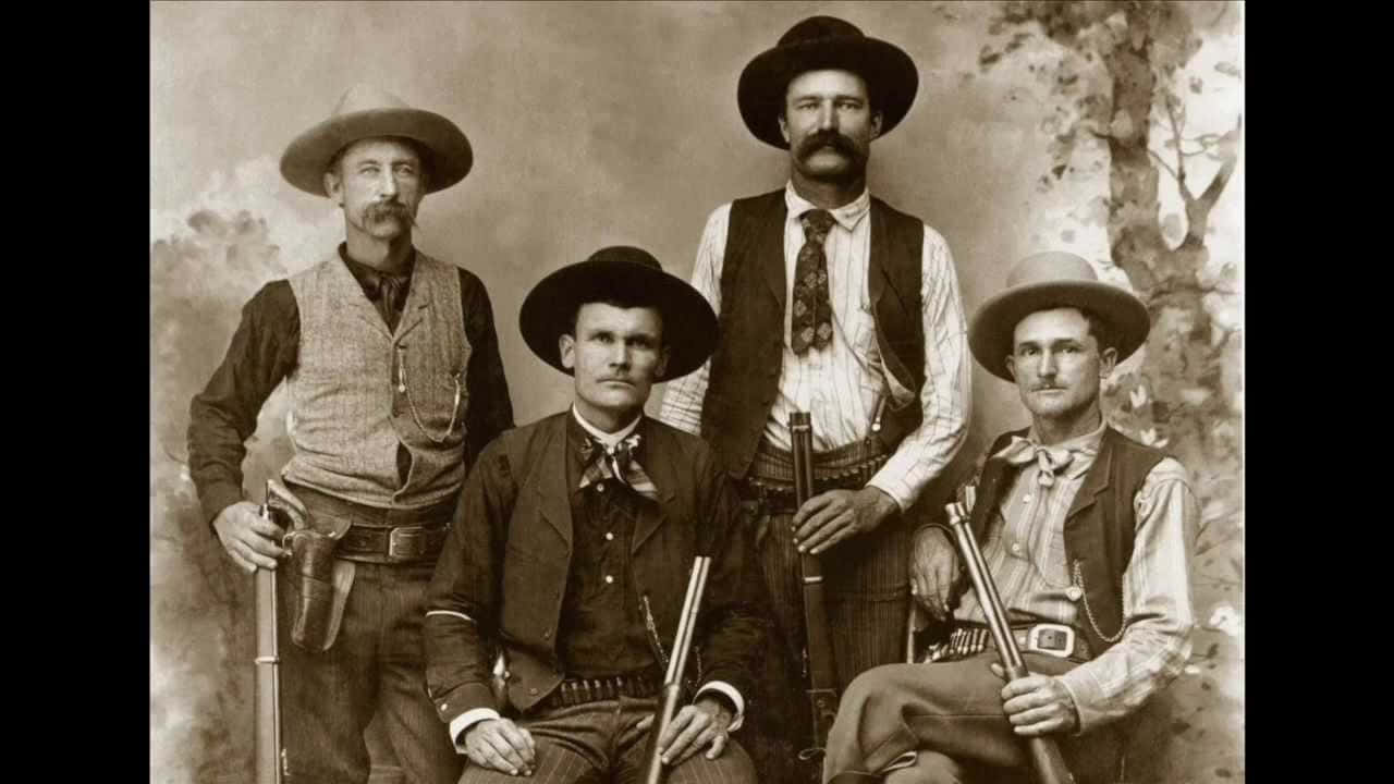 Old Western 桌布 壁紙 — 四位表情嚴肅的牛仔穿著復古服裝，在繪製的背景前拿著步槍，以棕褐色調的攝影棚肖像照姿勢擺拍。