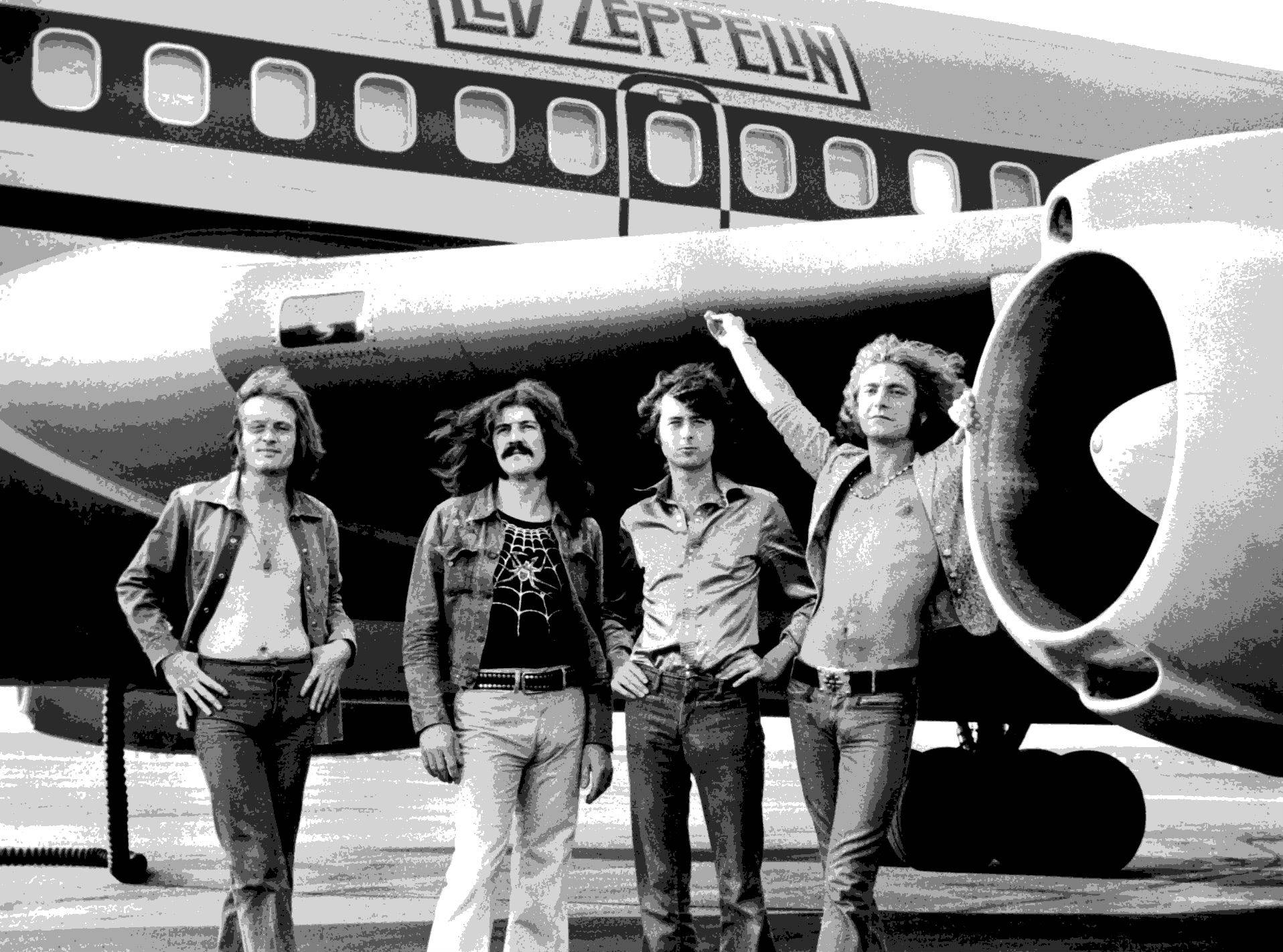 band 桌布 壁紙 — 四位身穿70年代服裝的男子自信地站在一架側面寫有「LED ZEPPELIN」的大型飛機前。