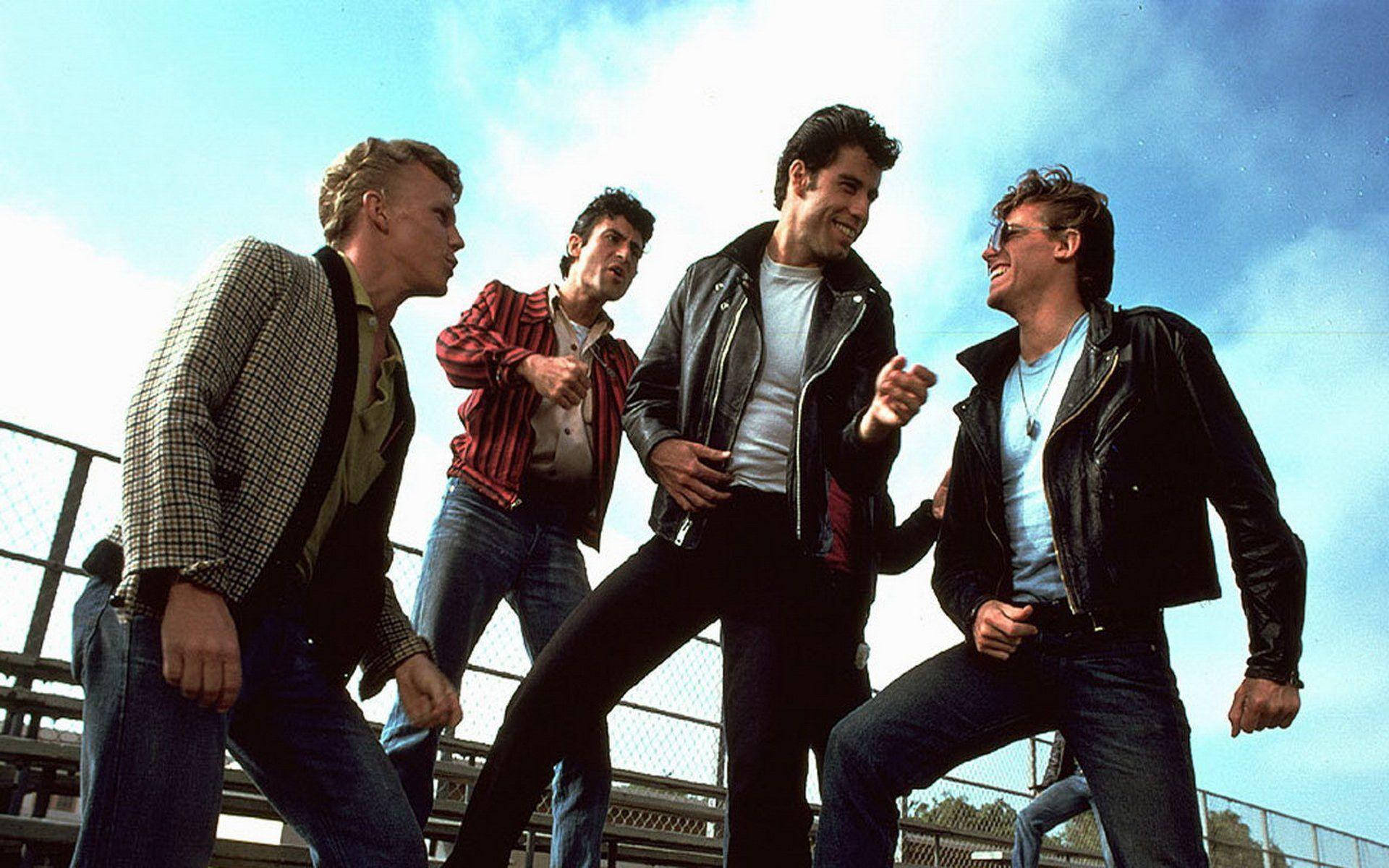 Grease 桌布 壁紙 — 四位身穿80年代風格皮夾克和牛仔褲的年輕男子，在明亮的藍天下於看台上充滿活力地擺姿勢。