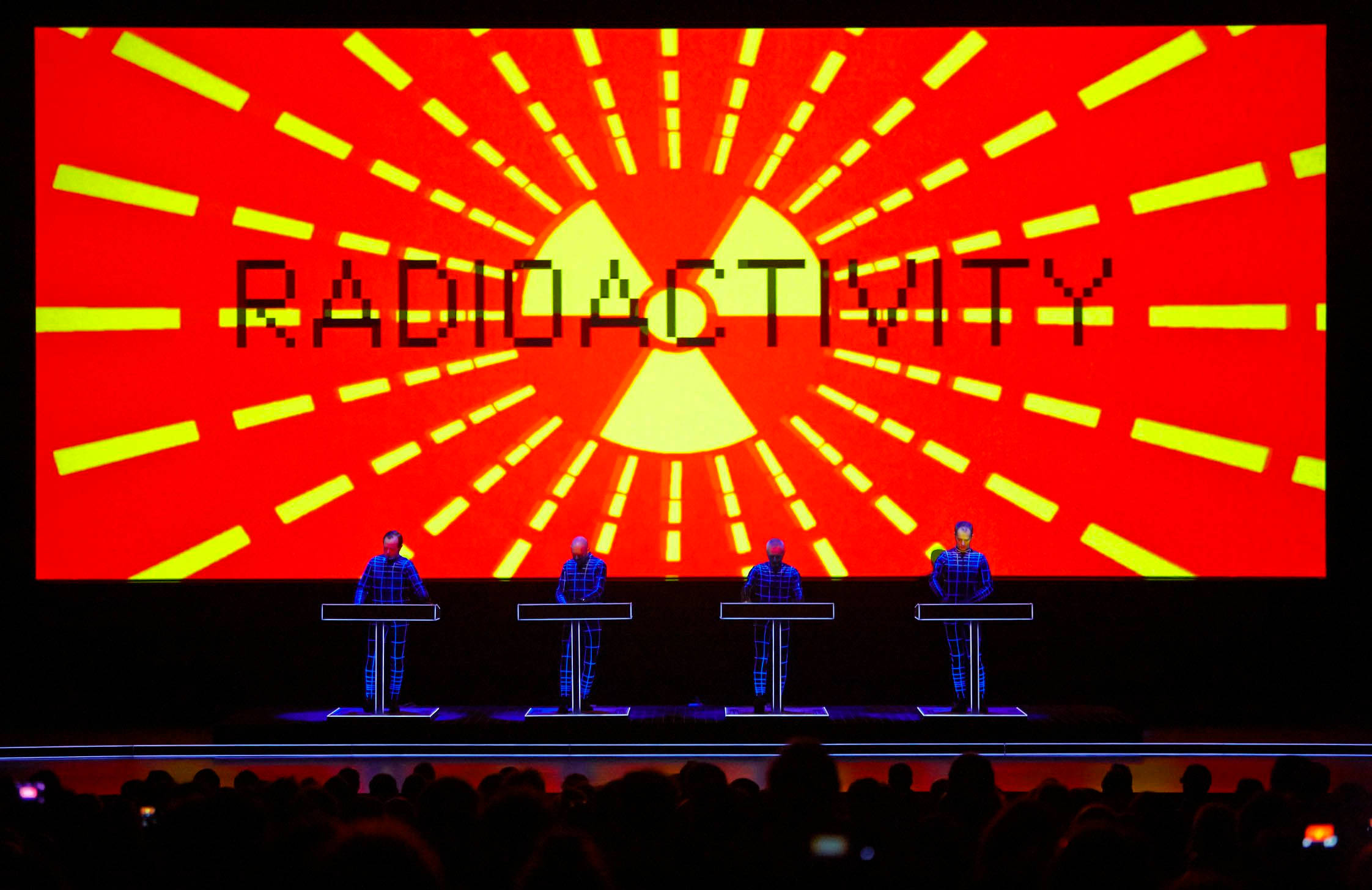 band 桌布 壁紙 — 四位身穿格紋西裝的音樂家在舞台上表演，舞台上方有一個巨大的紅色螢幕，顯示「RADIOACTIVITY」和一個發光的黃色輻射標誌。