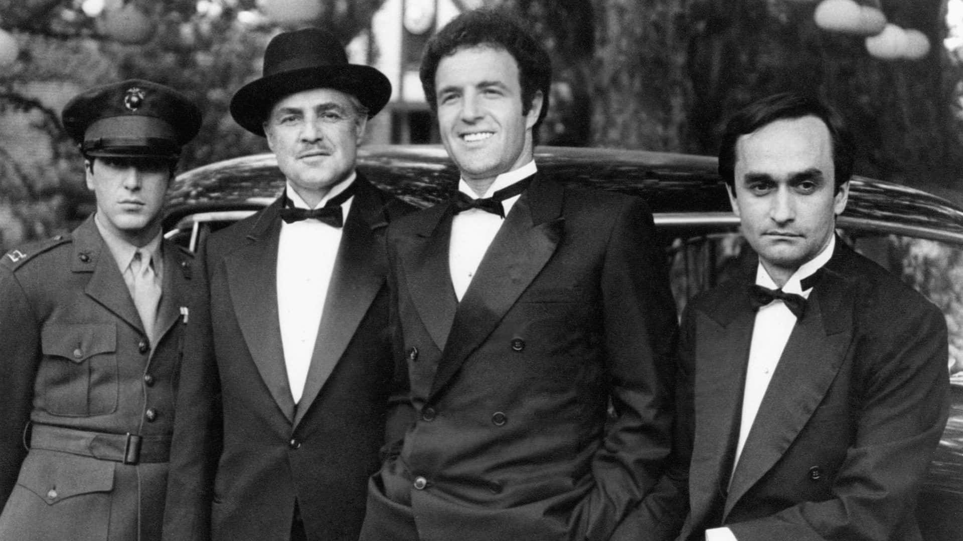 The Godfather 桌布 壁紙 — 四位身穿正式服裝和軍裝的男子在一張經典黑白照片中自信地站在一輛老爺車旁。