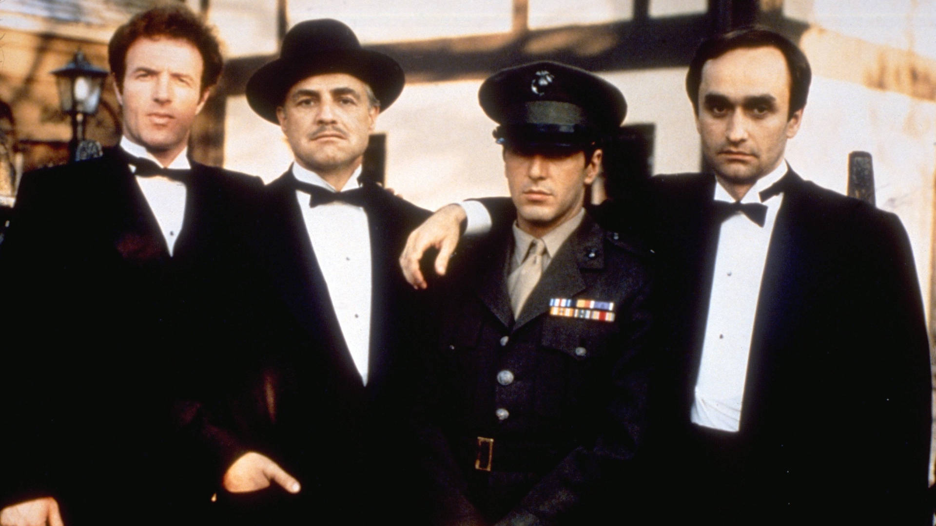 Godfather 桌布 壁紙 — 四位身穿正式黑色禮服的男人和一位身穿軍裝的男人，在陽光照射的建築物外牆前，表情嚴肅地站在一起。