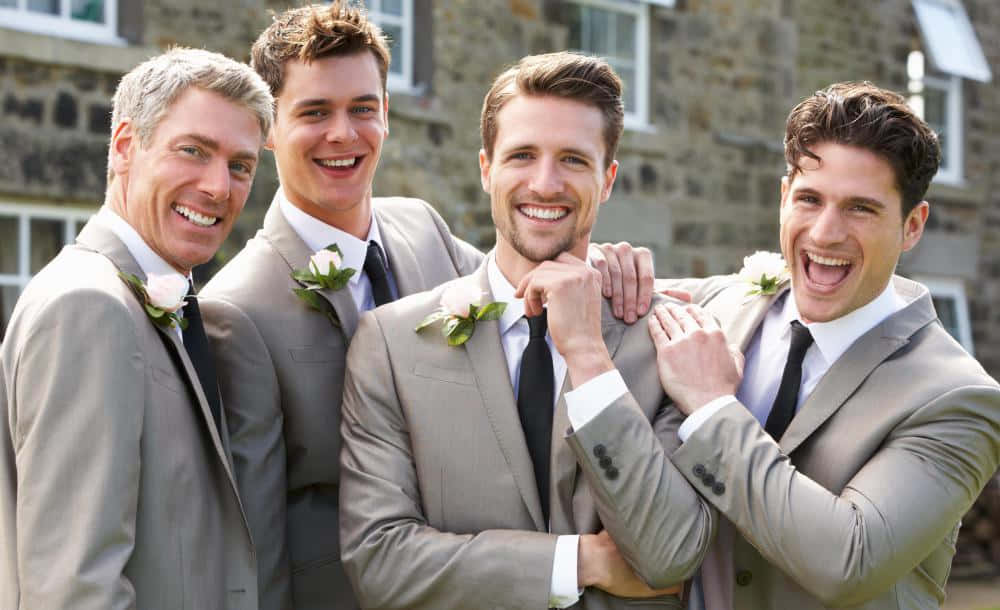 Groomsmen 桌布 壁紙 — 四位身穿灰色西裝的快樂伴郎，在石建築背景下，佩戴白色胸花一起大笑。