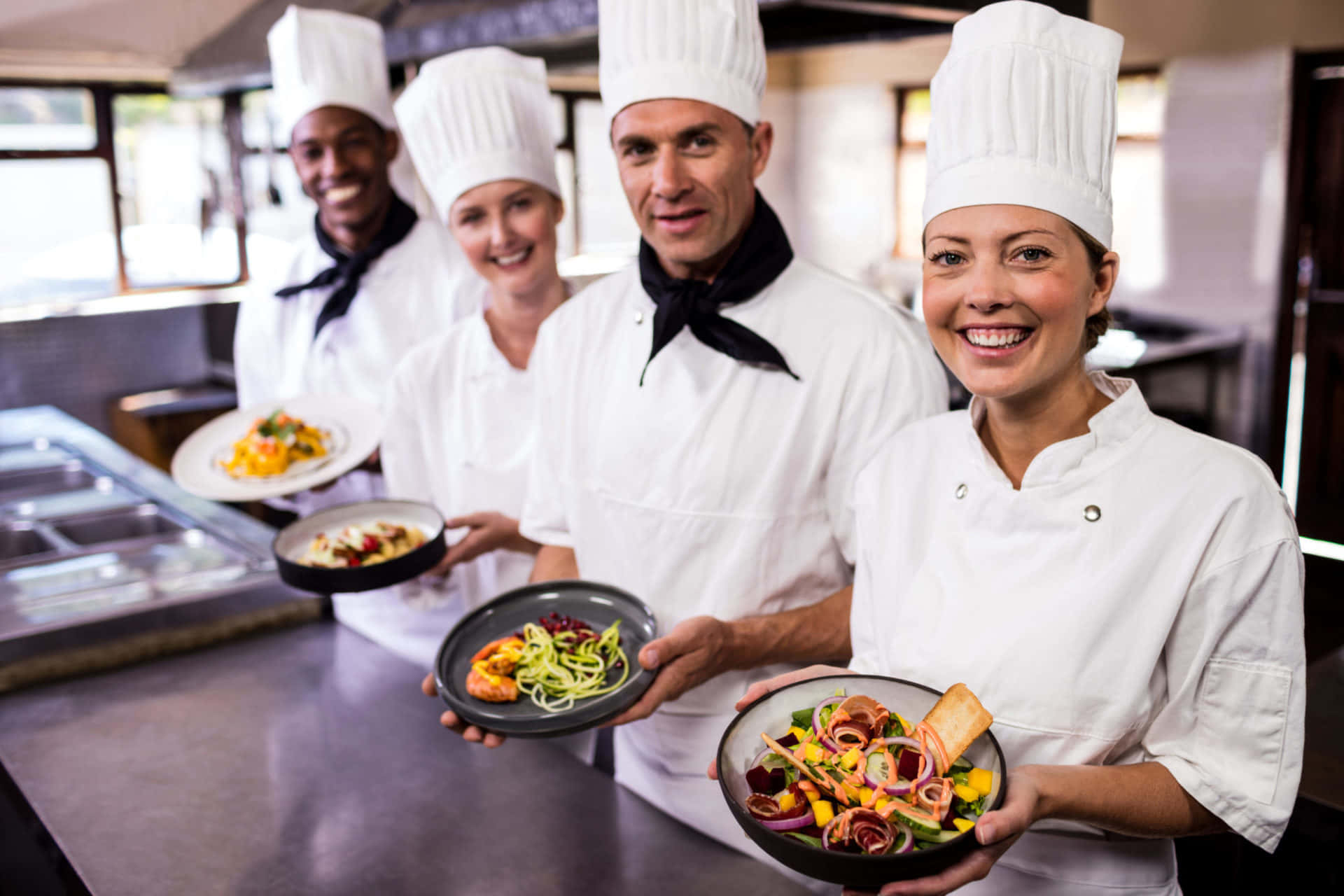 烹飪 桌布 壁紙 — 四位身穿白色廚師服的笑臉廚師，在明亮的專業廚房裡自豪地展示色彩繽紛的擺盤菜餚。