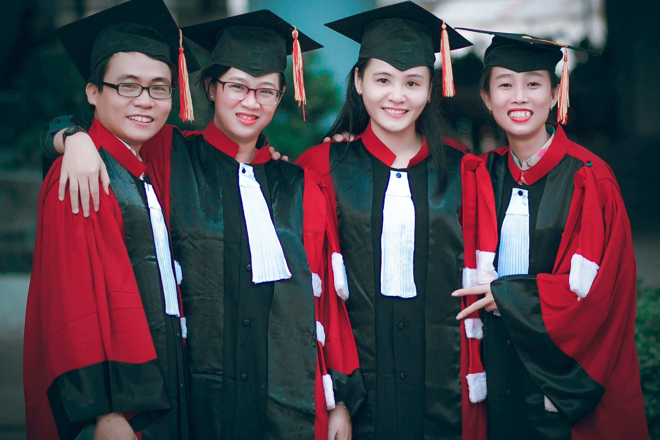 Graduation Ceremony 桌布 壁紙 — 四位身穿紅黑色畢業帽和袍的快樂畢業生，在戶外緊密地擺姿勢，為慶祝照片燦爛微笑。
