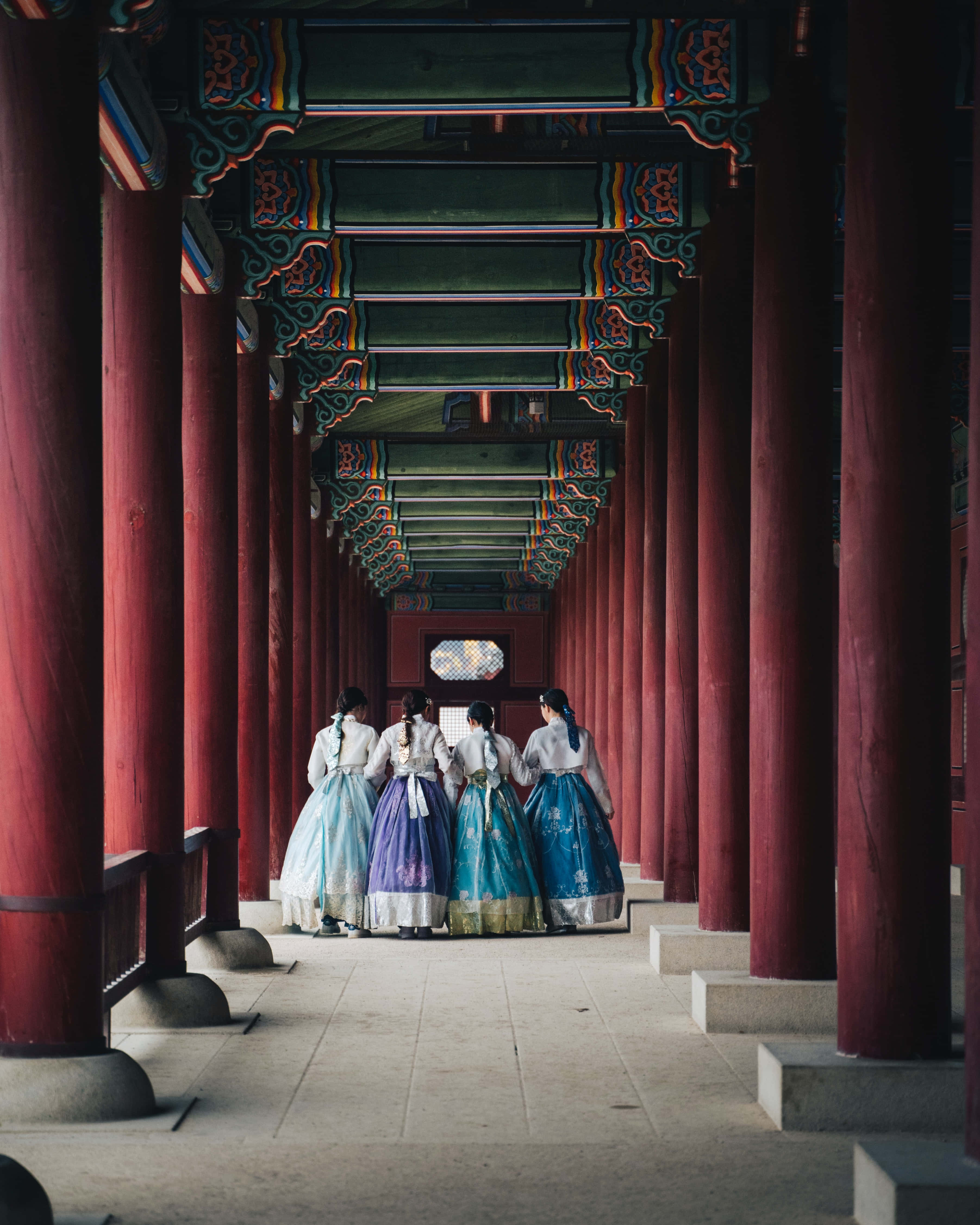 韓國 桌布 壁紙 — 四位身穿色彩繽紛韓服的女士，在由紅色柱子和華麗綠色天花板框住的傳統韓國寺廟走廊中走開。