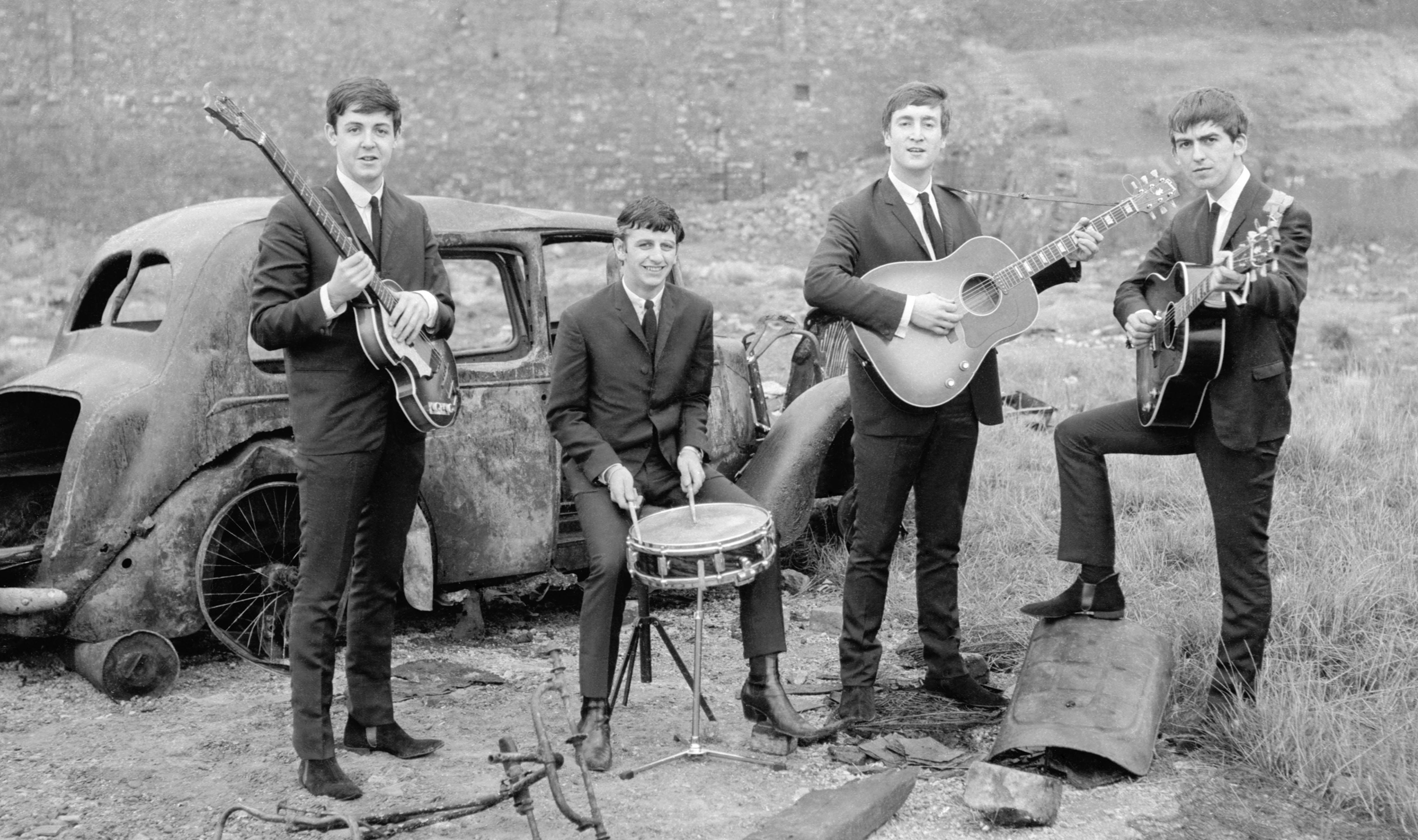 the beatles 桌布 壁紙 — 四位身穿西裝的年輕男子在草地上，在一輛生鏽的經典汽車旁彈吉他和打鼓。
