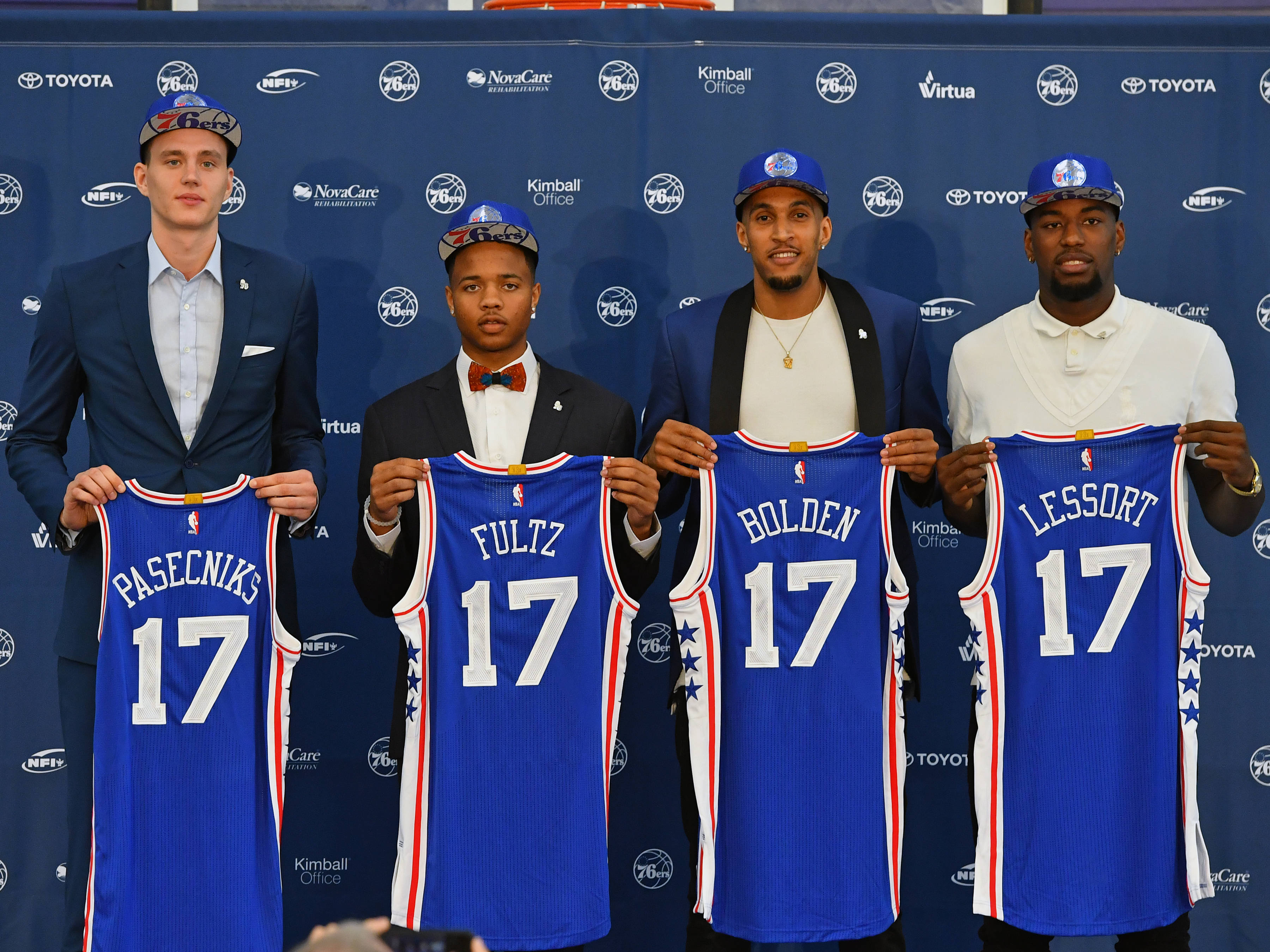 76ers 桌布 壁紙 — 四位身穿西裝的籃球運動員在品牌背景下手持帶有名字和17號的藍色費城76人球衣。